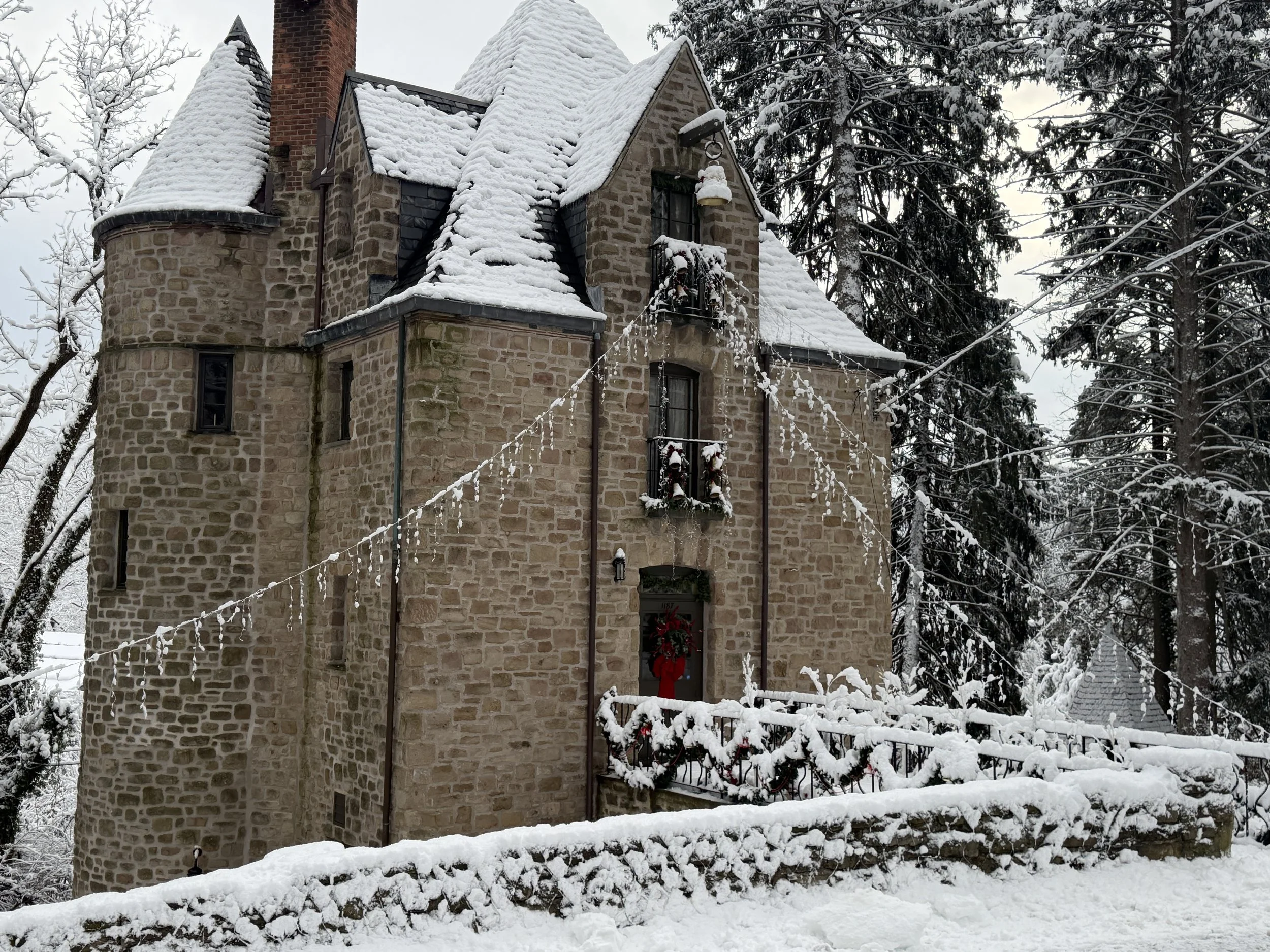 A stone castle-style house with snow-covered roof and yard, decorated for Christmas with wreaths, garlands, and a red bow on the door, amid snow-covered trees.