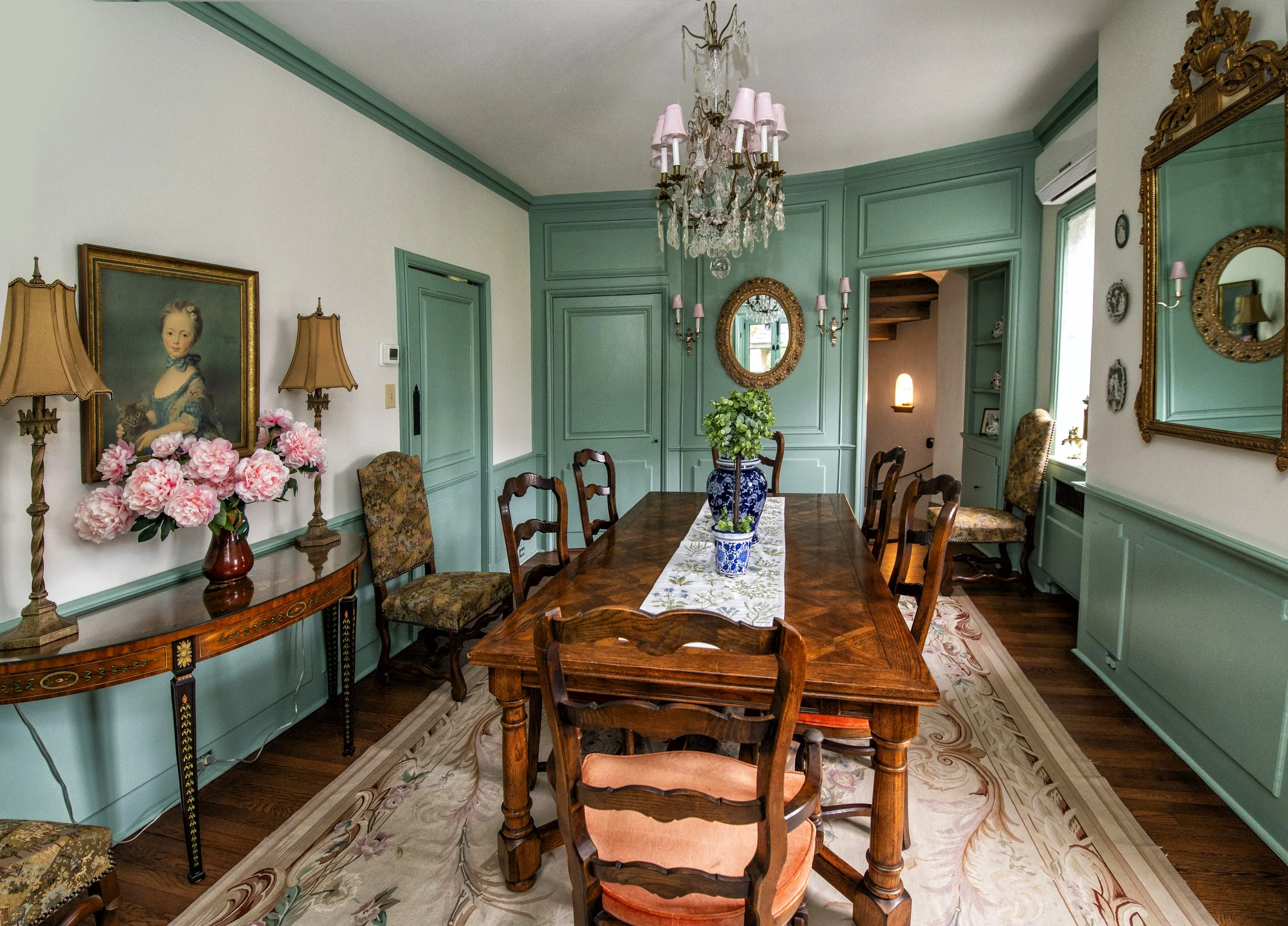 A vintage dining room with teal walls, a wooden dining table with a white table runner and potted plants, floral upholstered chairs, pink lampshades on wall sconces, a chandelier, a floral rug, and framed paintings and mirrors on the walls.