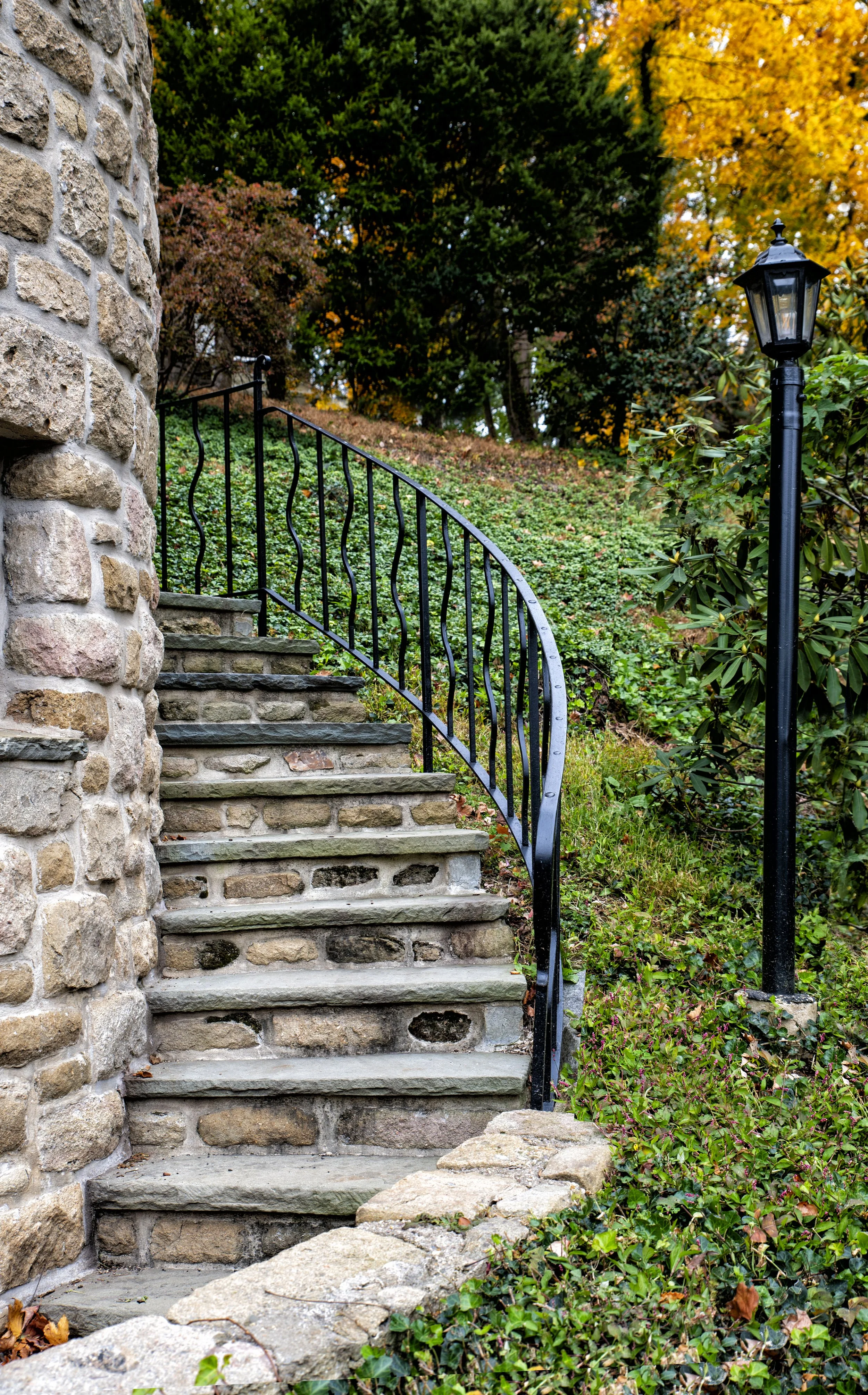 Stone steps with black metal railing leading up a garden hillside, with a vintage-style lamp post on the right, surrounded by green and autumn-colored trees.