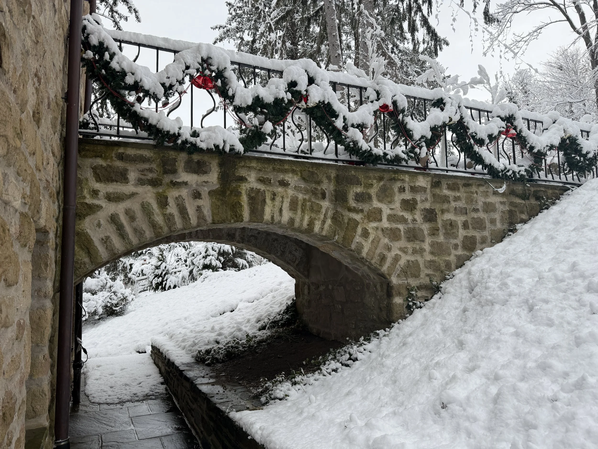Snow-covered stone bridge with black wrought iron railing decorated with Christmas garland and red ornaments, over a snow-covered pathway.