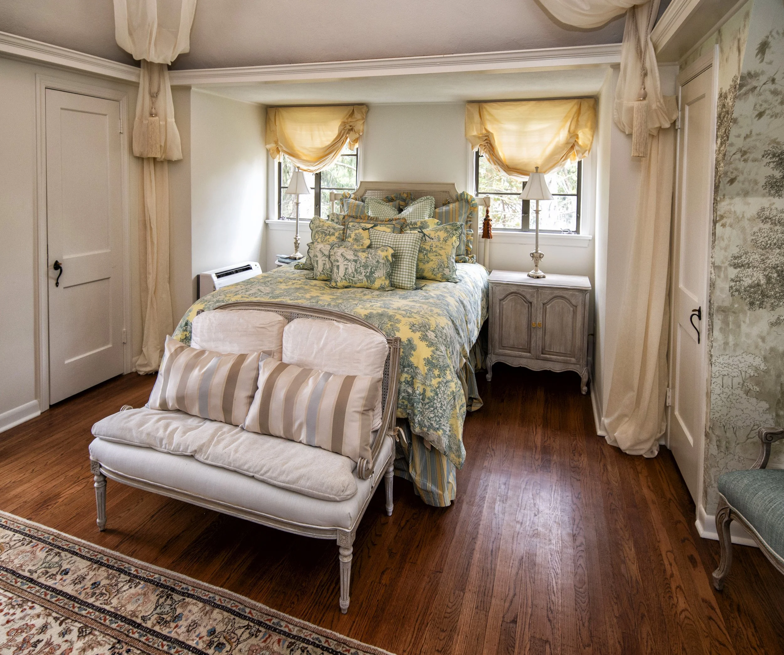 A cozy vintage bedroom with a bed adorned with floral and gingham throw pillows, bedside tables with lamps, a window with yellow curtains, an upholstered bench at the foot of the bed, and hardwood floors.