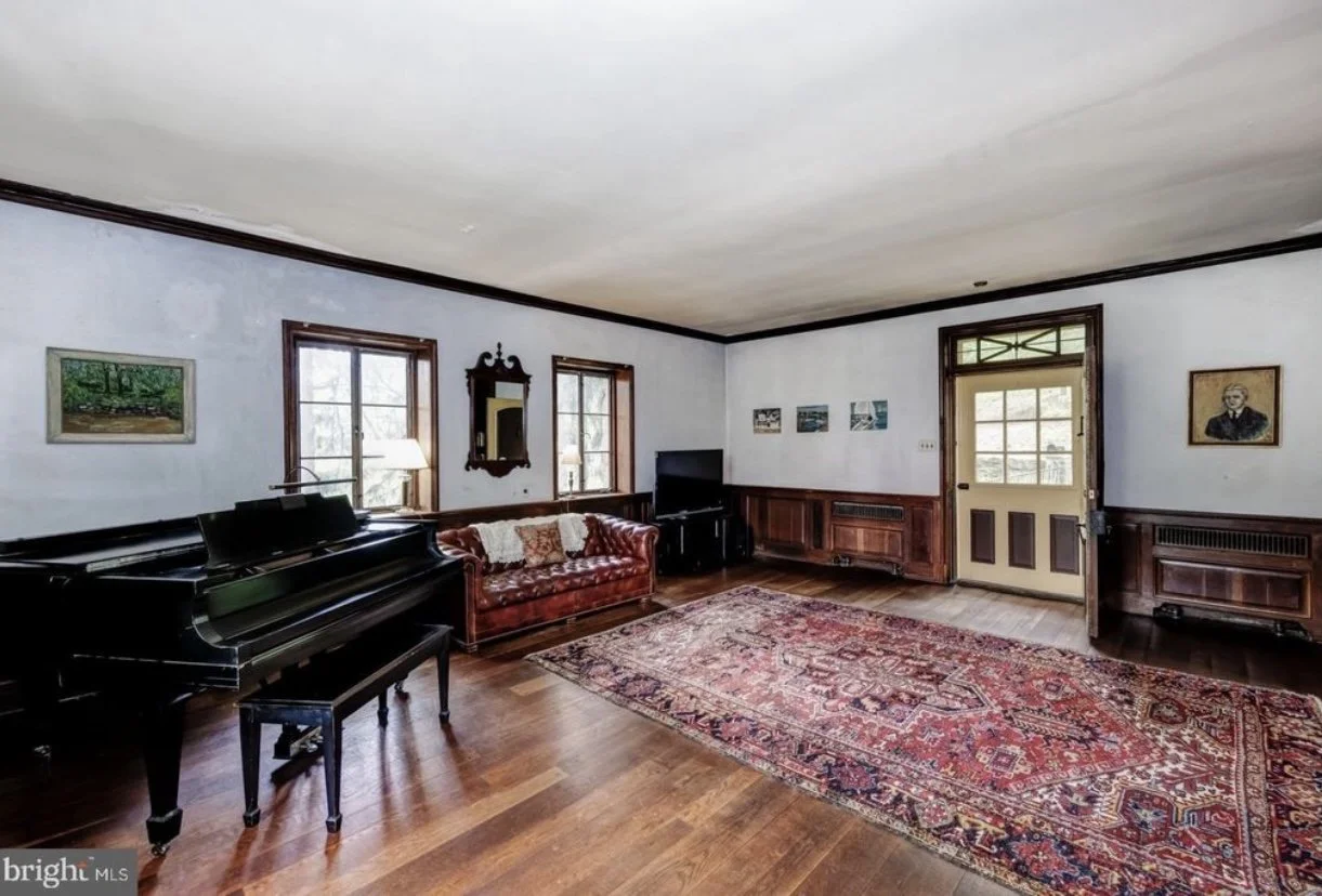 Living room with a piano, a red leather couch, a television, hardwood floors, and a large area rug, with wooden trim around the windows and door.
