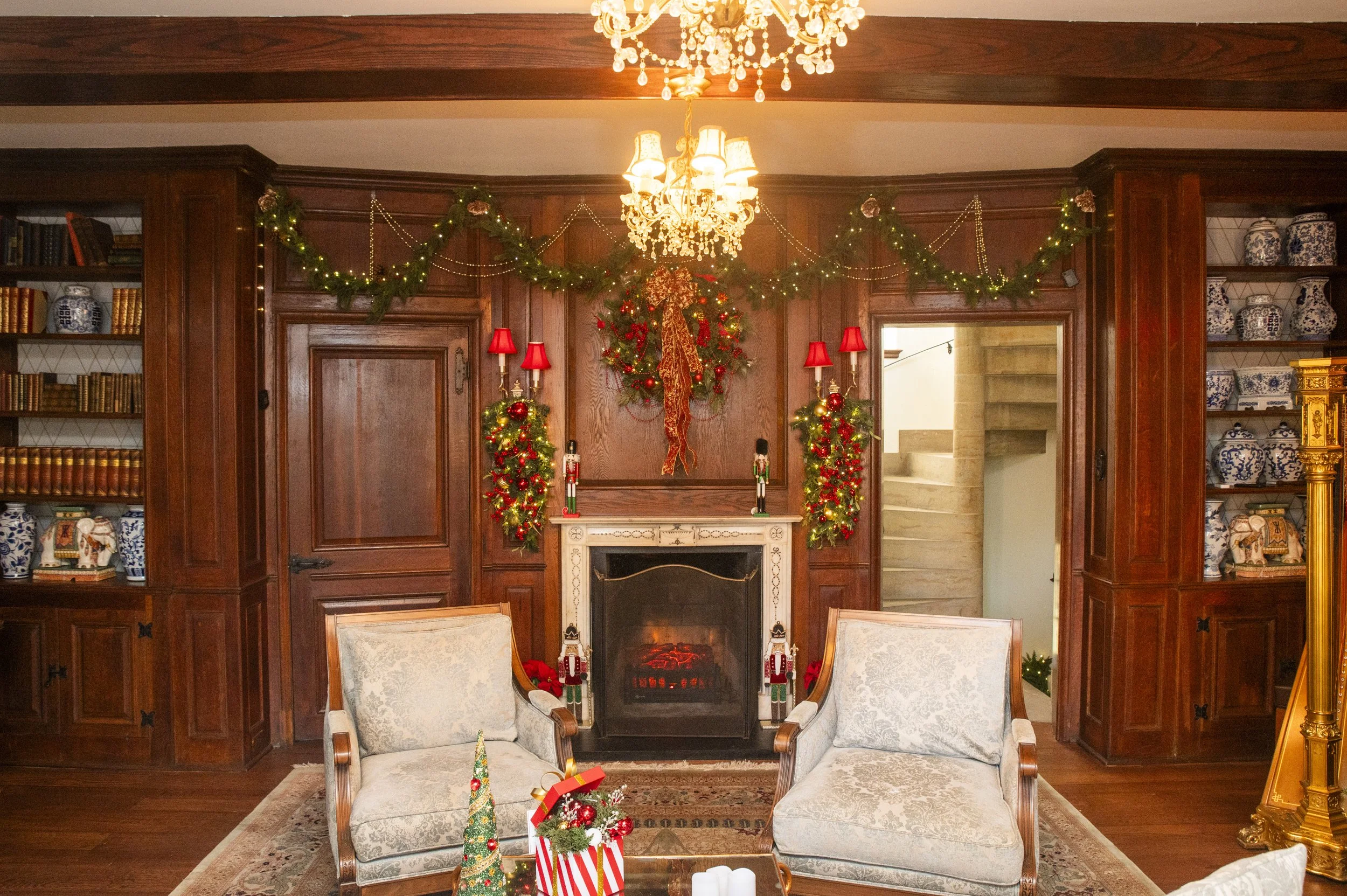 A cozy living room decorated for Christmas. There is a fireplace with garlands, nutcracker dolls, and red lamps. A chandelier hangs from the ceiling. Two armchairs are in front of the fireplace with holiday decorations on a table nearby.