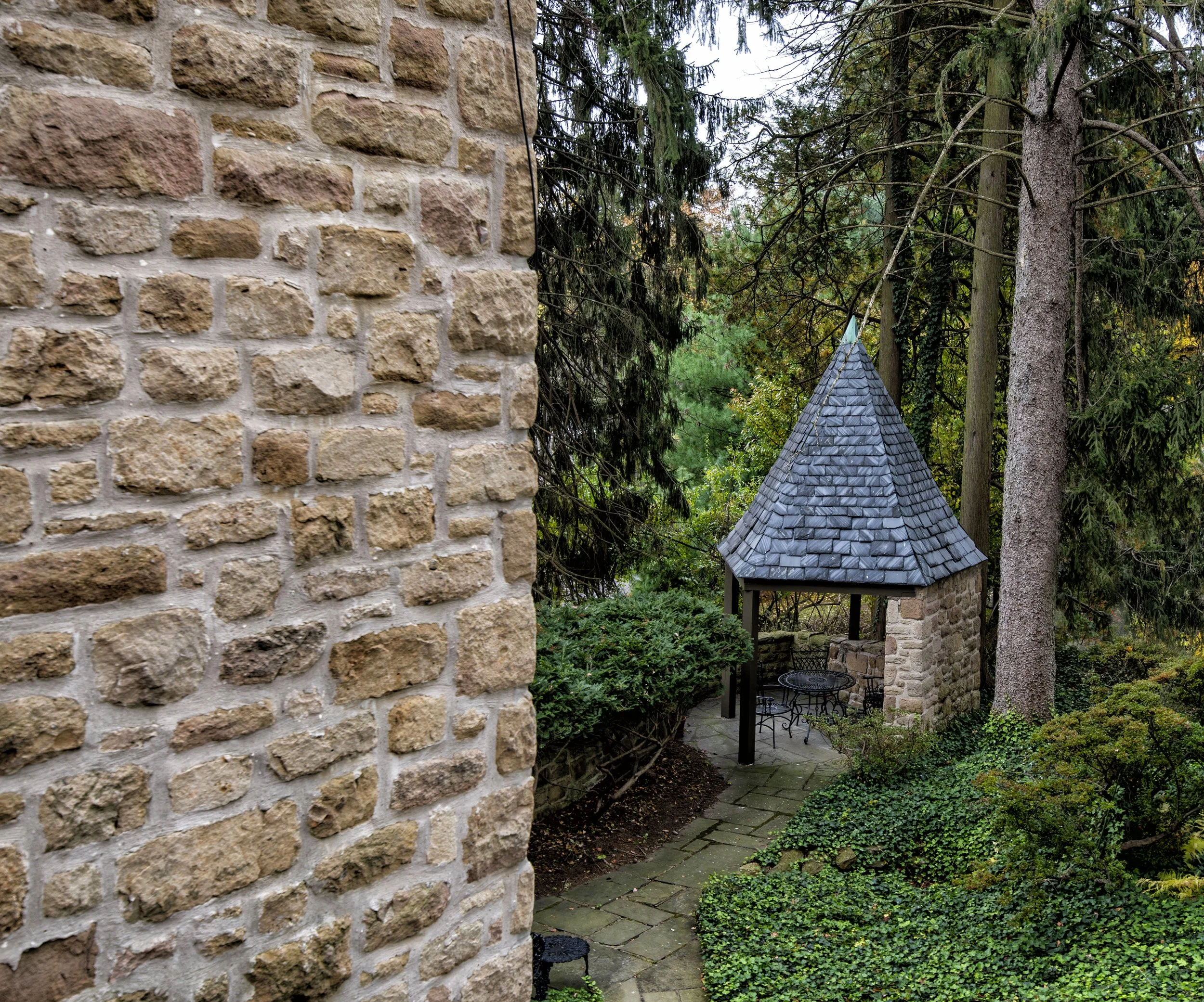 A garden with a stone pathway leading to a small stone and wood gazebo with a conical slate roof, surrounded by trees and green bushes.
