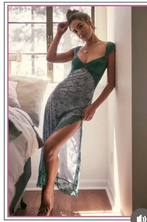 Young woman standing in a narrow space by the window, wearing a lace dress, posing with one hand on her head and the other on her thigh.