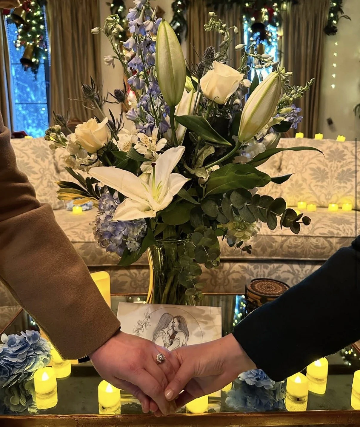 A floral arrangement with white lilies, roses, and blue flowers in a glass vase placed on a table with yellow candles around it. Two people are holding hands in front of the arrangement.