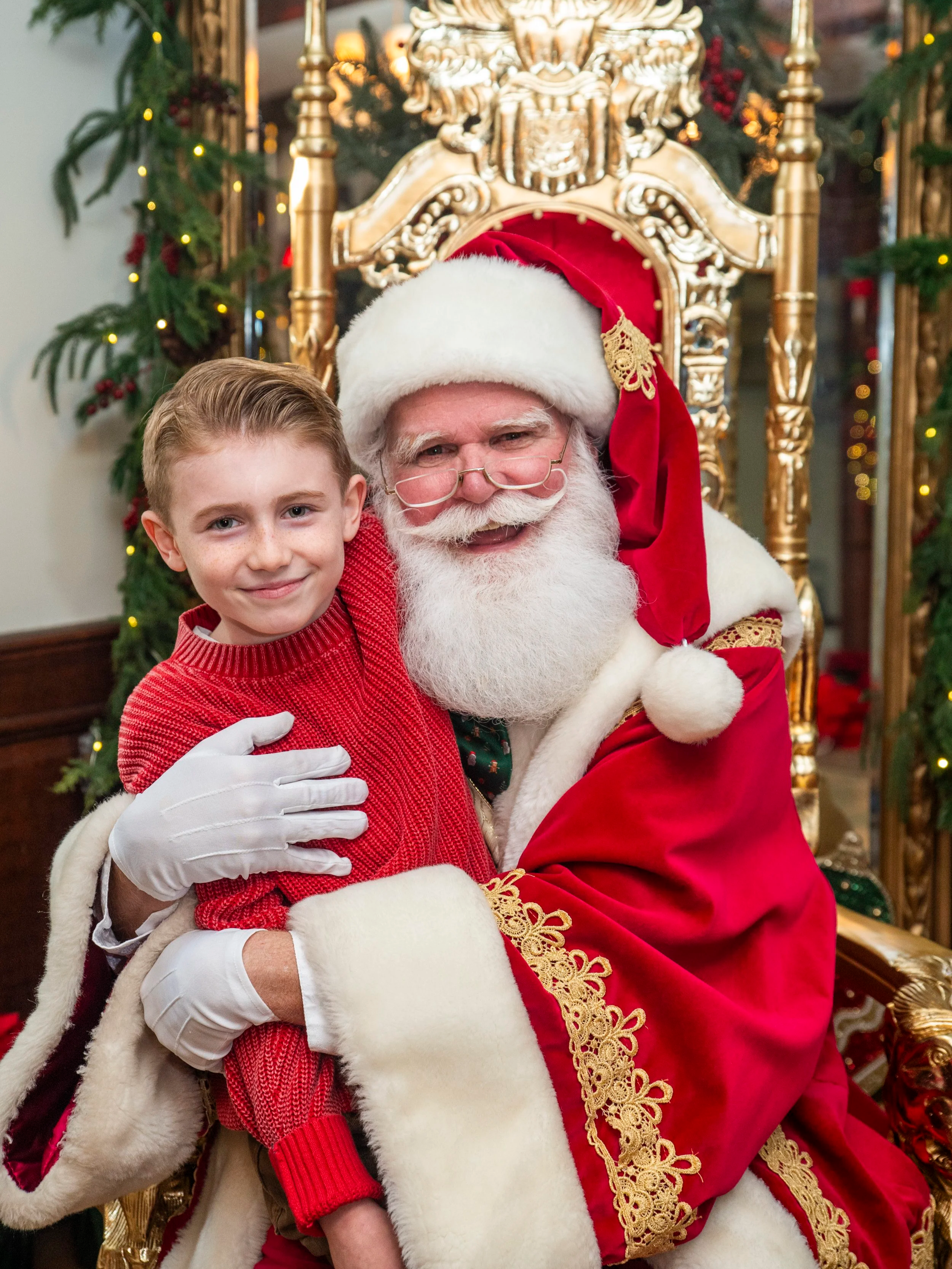 A young boy sitting on Santa Claus's lap, both smiling. Santa is dressed in a traditional red and white suit with gold embroidery, sitting on a decorated gold throne with Christmas greenery and lights in the background.