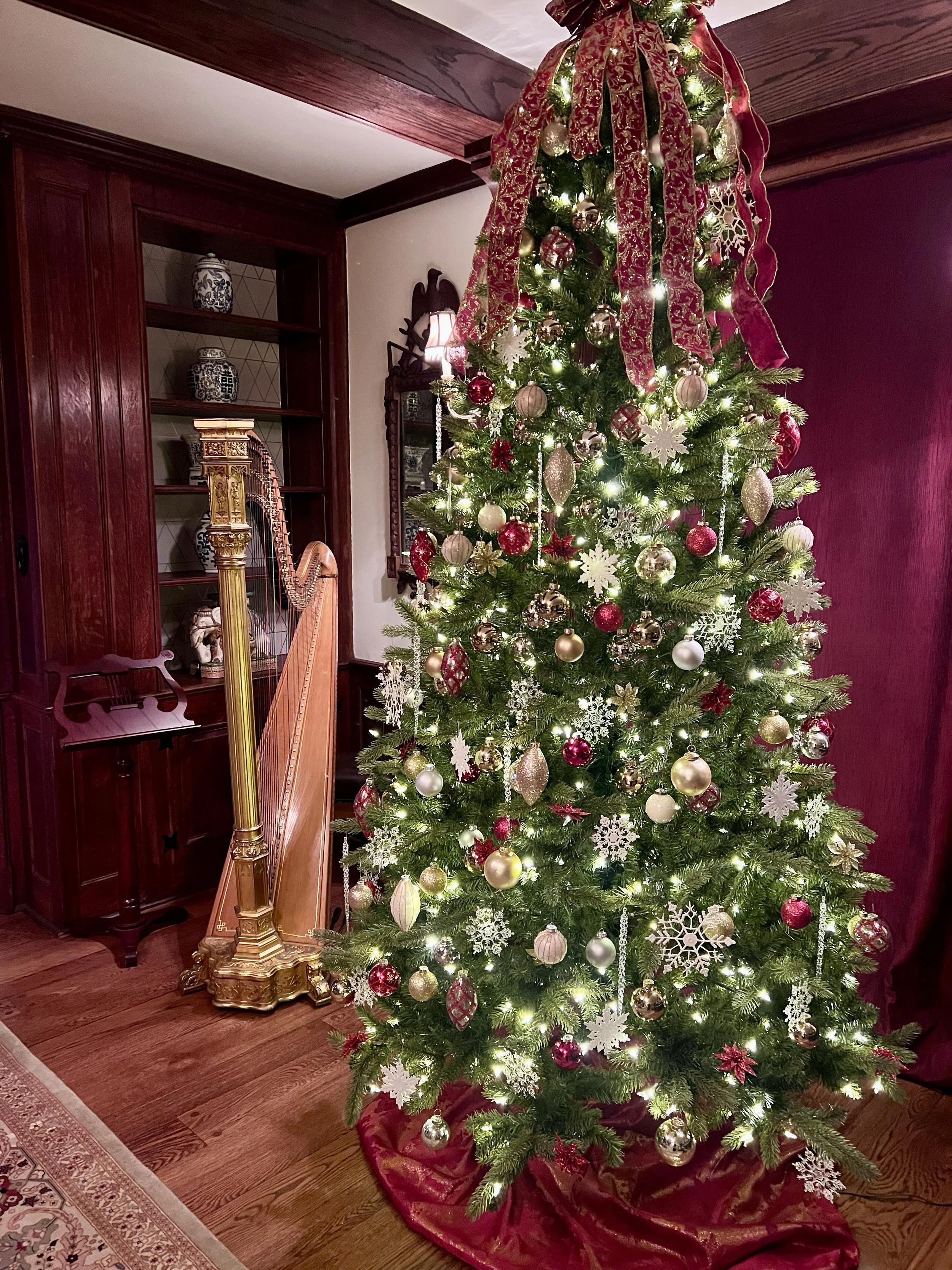 Decorated Christmas tree with white, red, and gold ornaments, ribbons, and lights in a living room with wooden furniture and a harp.
