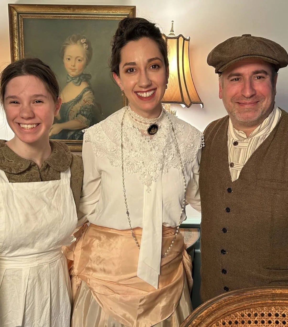 Three people dressed in historical clothing from the 19th century, standing in a warmly lit room with a portrait of a young girl in period dress hanging behind them.