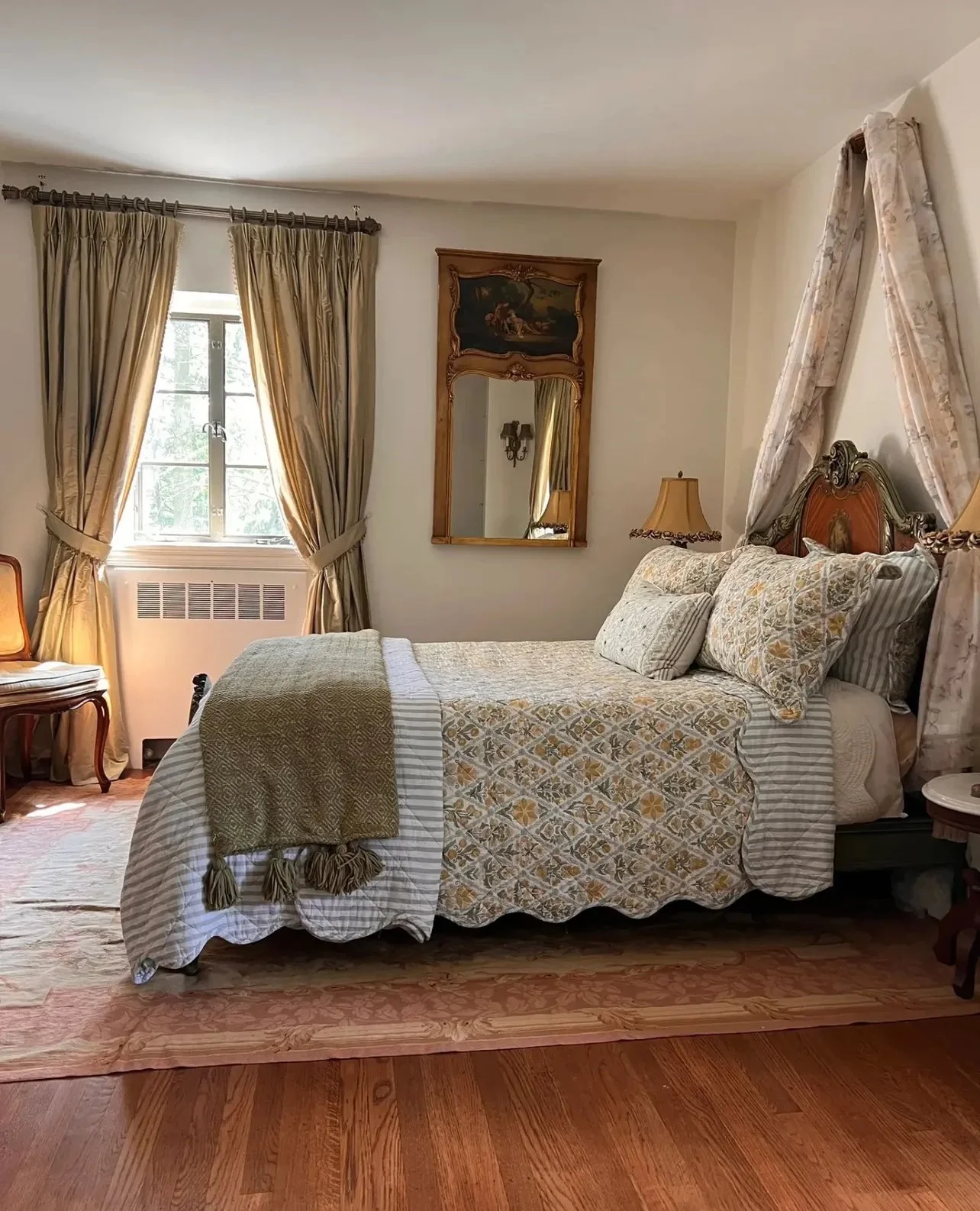A bedroom with a bed covered in a patterned quilt, with several pillows, beside a nightstand with a lamp, a window with gold curtains, an ornate mirror and wall sconces, and a chair near the window. The room has wooden floors and a vintage decor styl