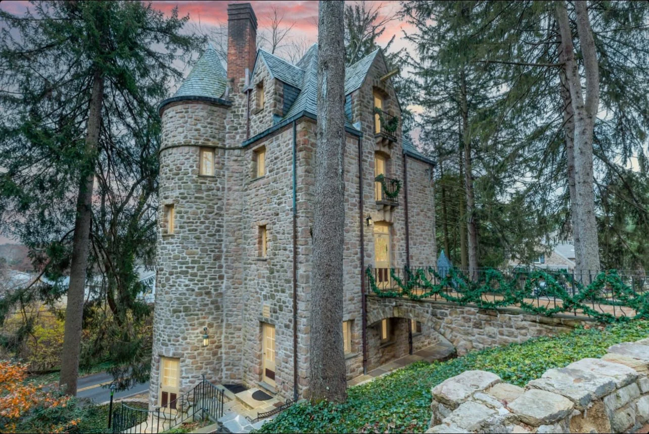 A stone castle-style house surrounded by tall trees, with warm lights glowing inside, and festive garlands decorating the balcony. The scene is at dusk with a pinkish sky.
