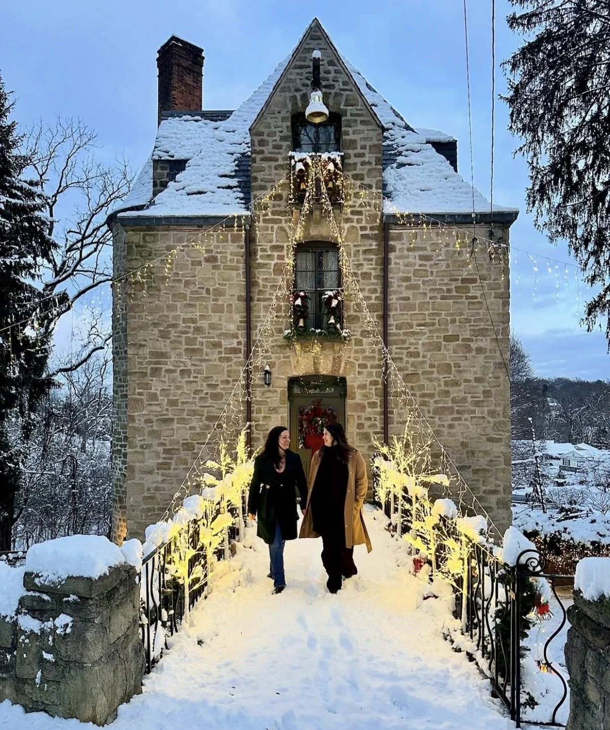 Two women walking across a snow-covered bridge decorated with festive holiday lights and wreaths toward a stone house with Christmas decorations on the windows and door, in a snowy winter landscape.