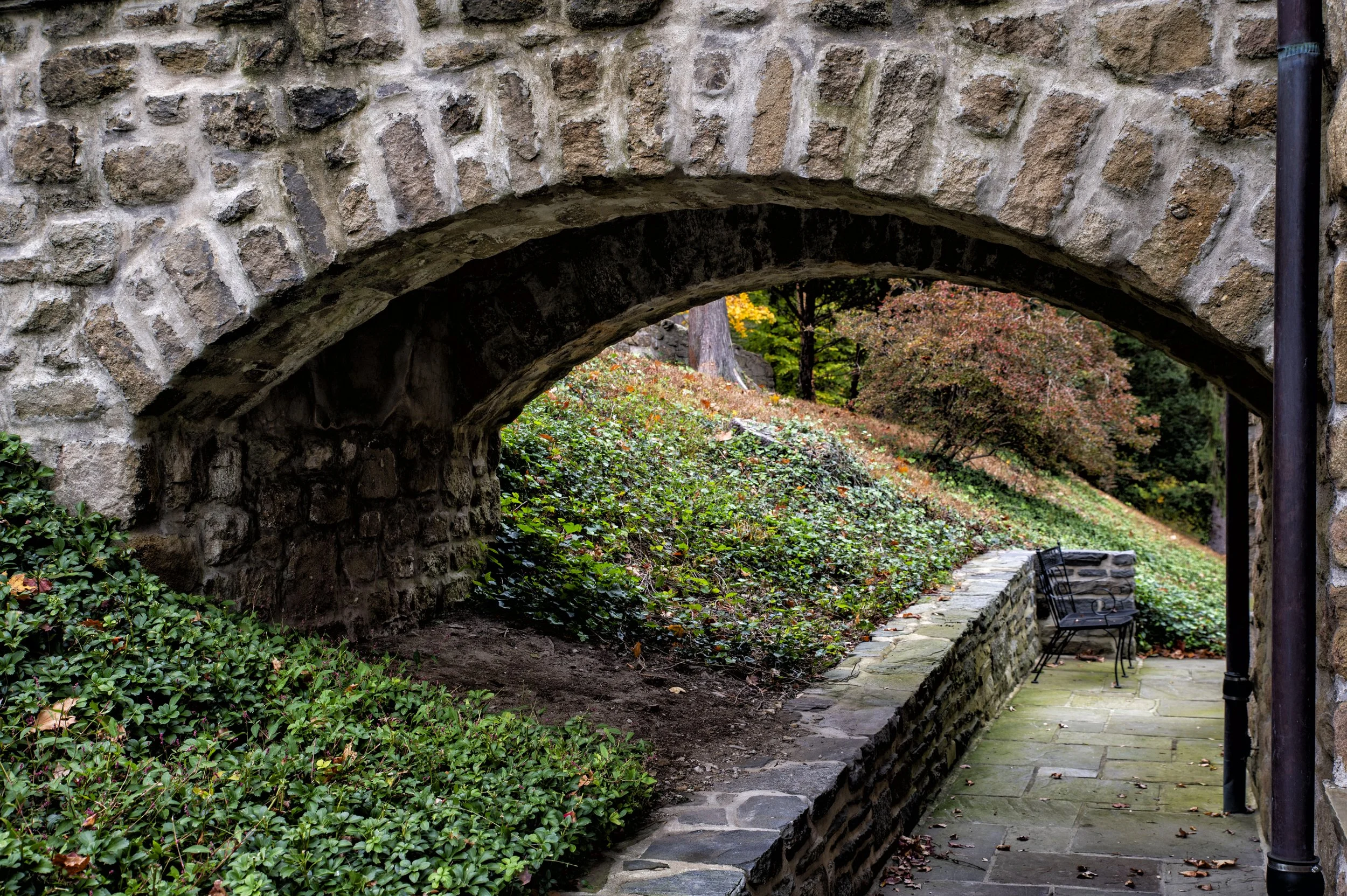 A stone archway leading to a garden with bushes, trees, and a paved patio with two black benches.