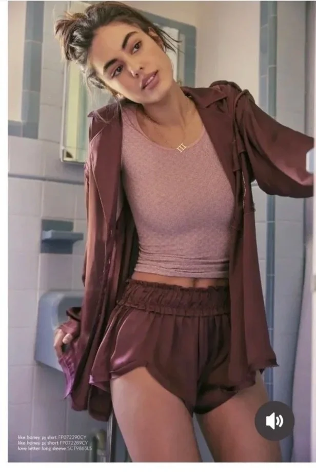 A young woman in a public restroom takes a selfie. She wears a light brown crop top, matching shorts, and a brown jacket. Her hair is tied up, and she is standing in front of a mirror.