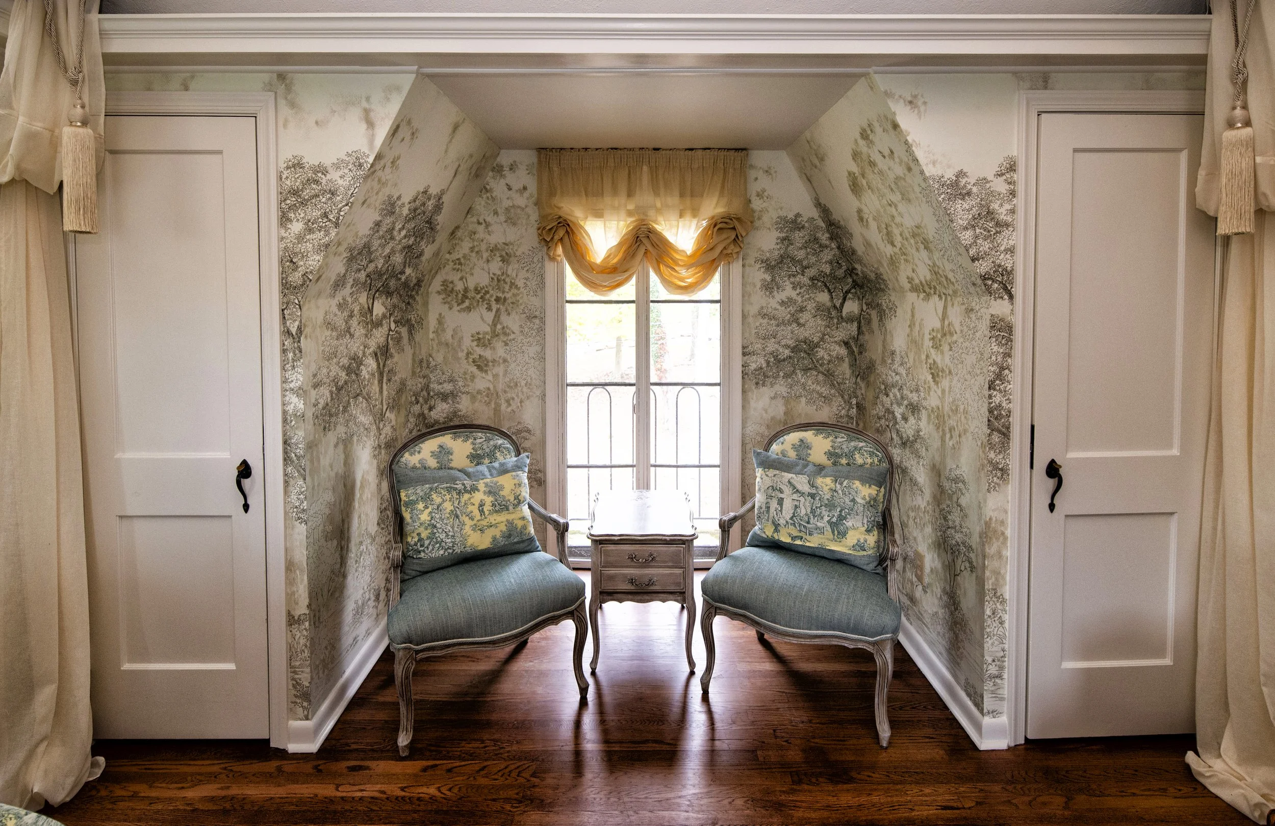 A cozy sitting nook with two vintage armchairs facing each other, small side table between them, and a window with sheer curtains and a yellow swag valance, inside a room with wallpaper depicting a forest scene and hardwood floors.