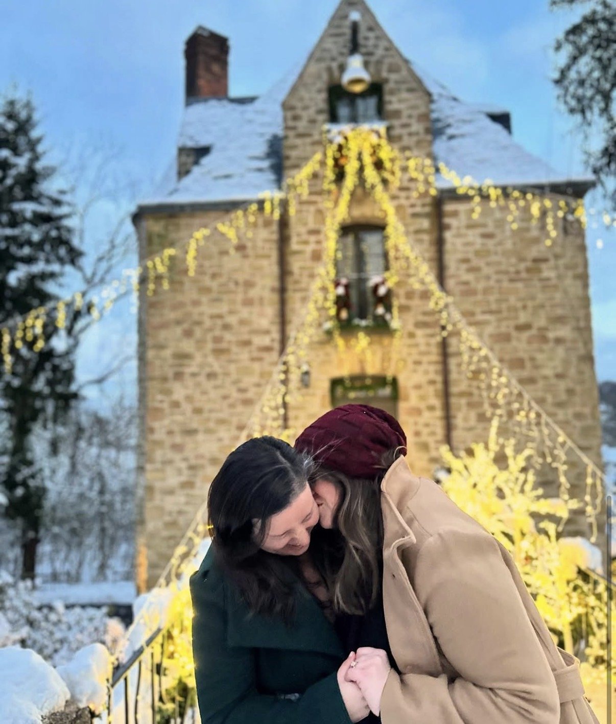Two women embrace and kiss in front of a snowy house decorated with yellow Christmas lights and wreaths during evening time.