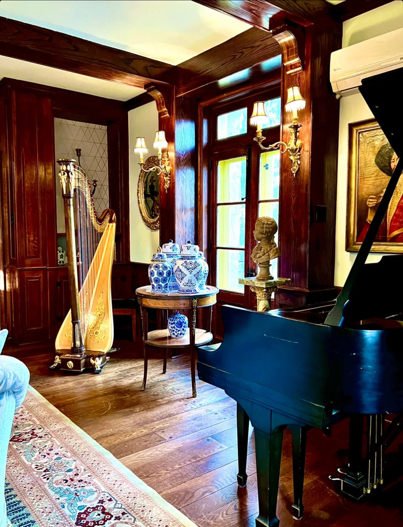 A cozy, elegant room with wooden walls and floors, decorated with antique furniture, a black grand piano, blue and white porcelain vases on a small table, and a golden bust on a pedestal. There are two wall-mounted lamps beside a large window, and a 
