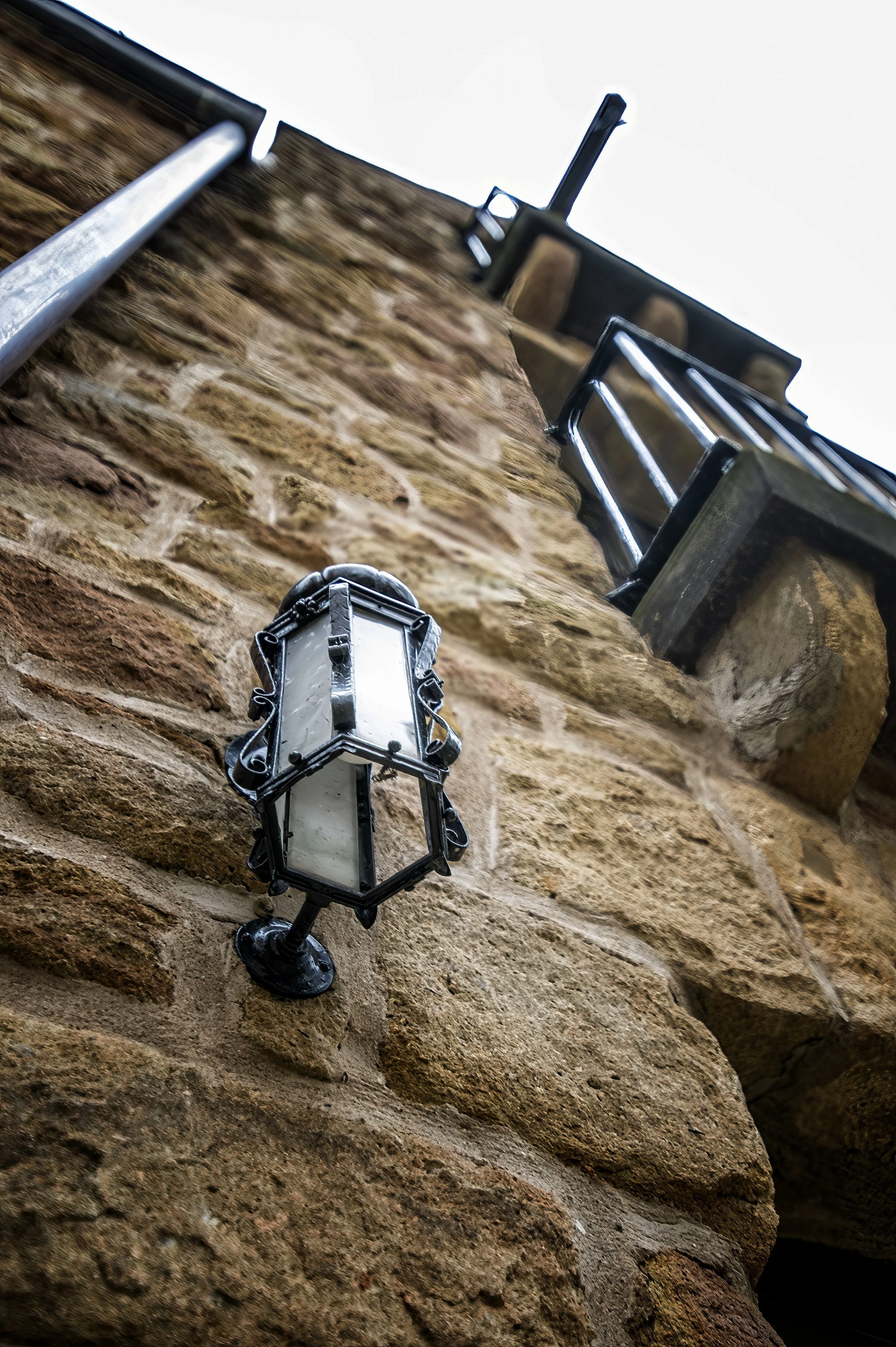 Close-up of a wall-mounted lantern with a rustic design, attached to a brick wall beneath a wooden balcony with metal railings, taken from below at an upward angle.