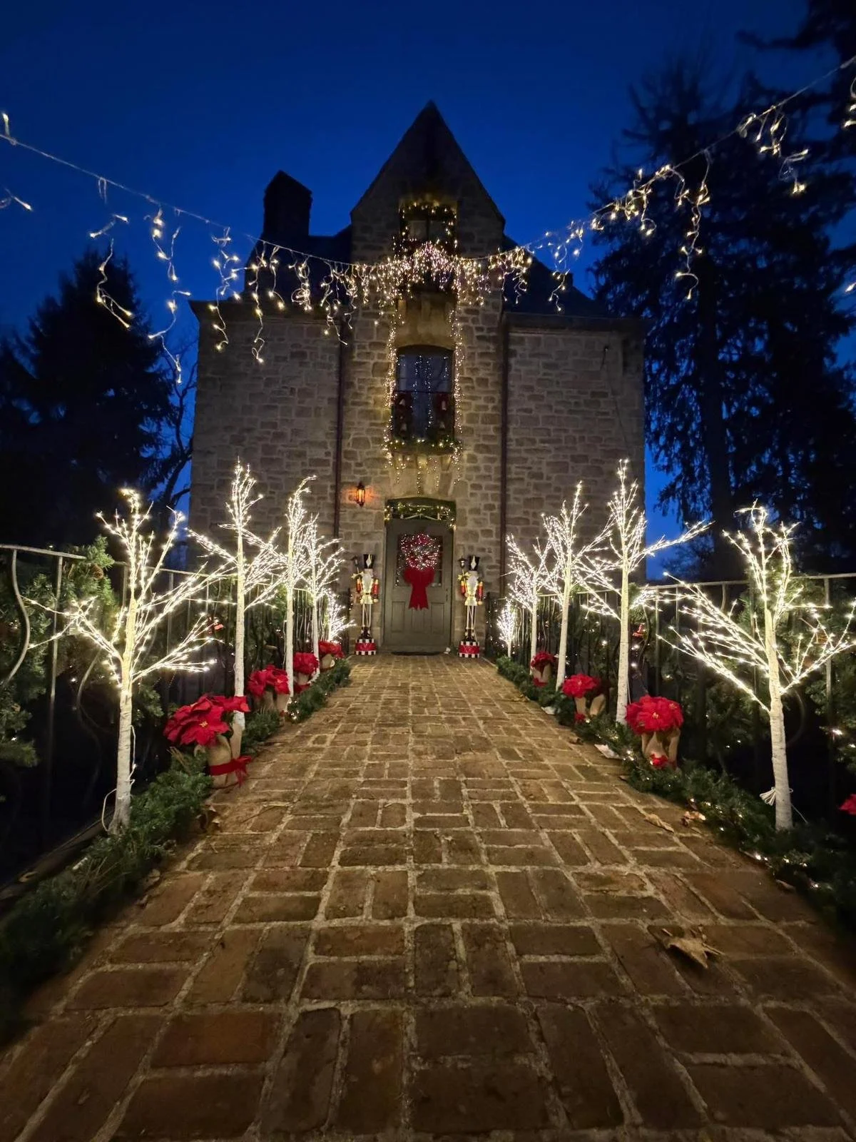 A brick pathway leading to a stone house decorated for Christmas with string lights, wreaths, and poinsettias, surrounded by trees.