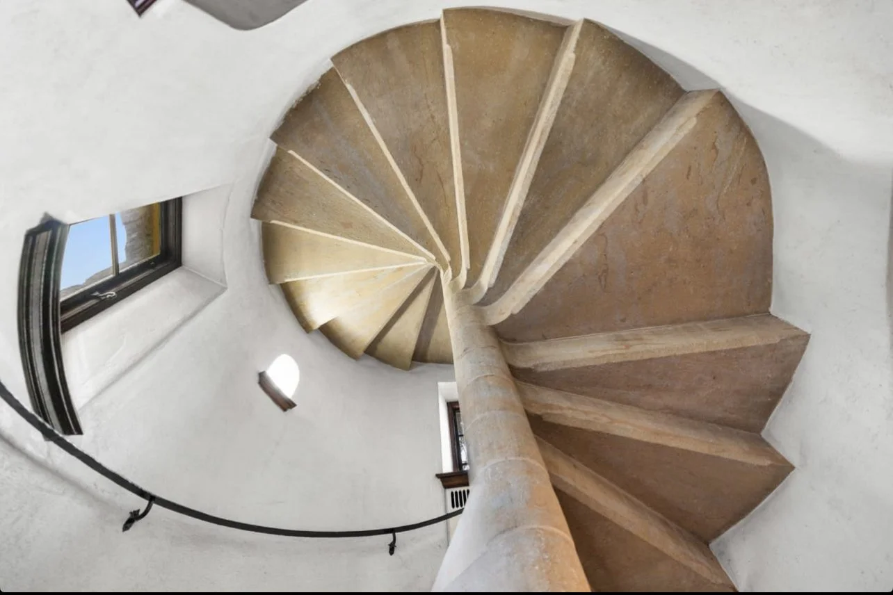 Interior view of a spiral staircase made of beige stone with a cylindrical central support and small windows on the white walls.