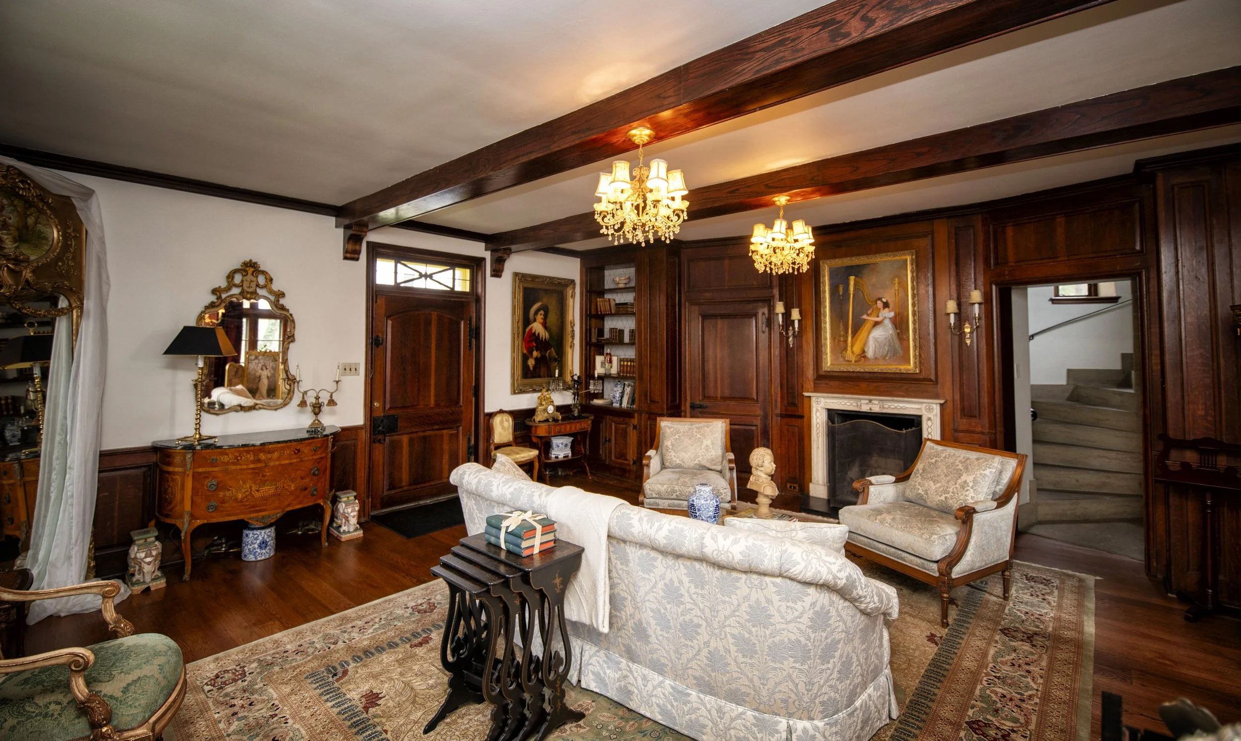 Living room with wood-paneled walls, ornate furniture, chandelier lighting, and staircase in the background.