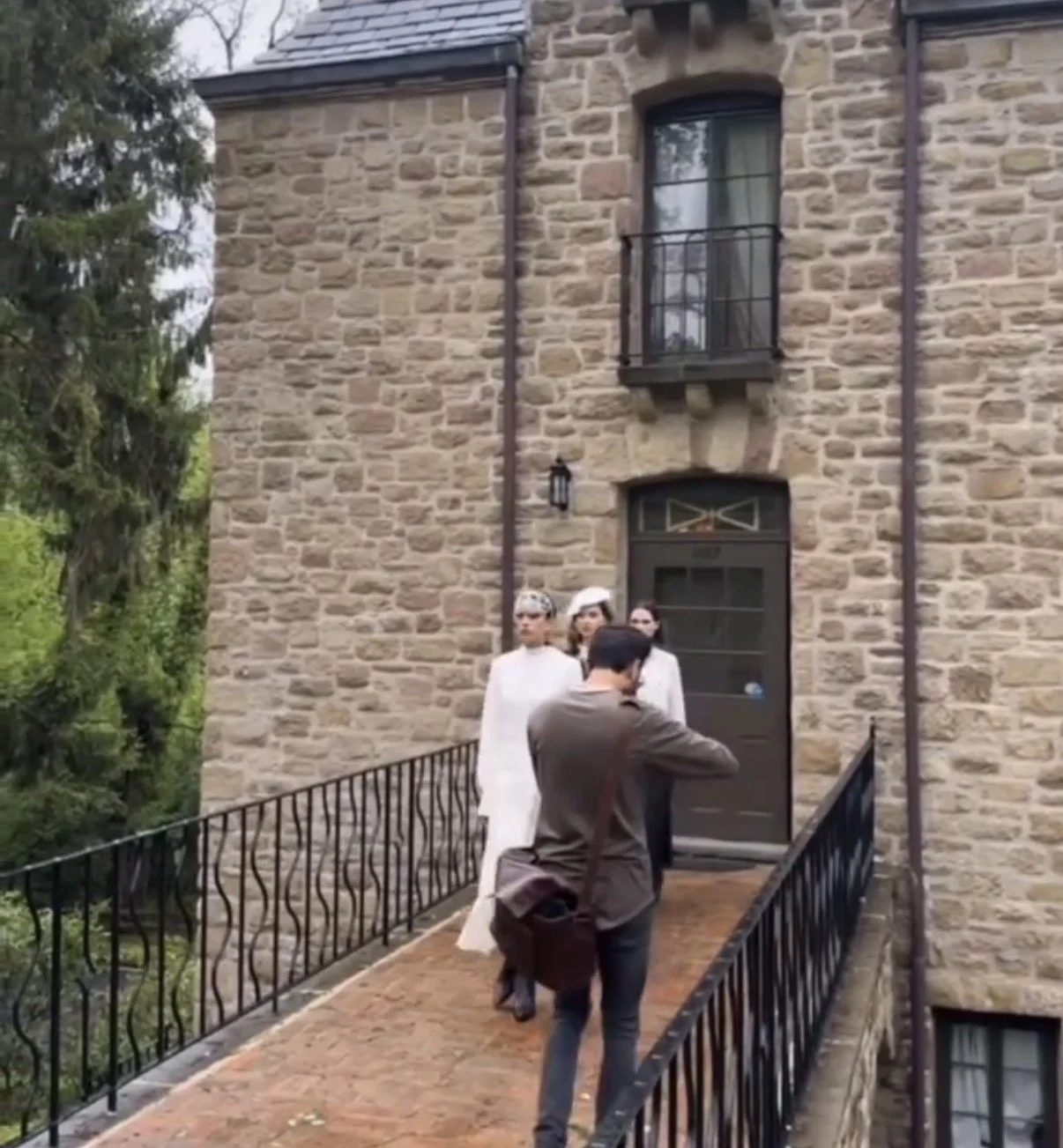 Photograph of three women standing outside a stone building, with a person taking their picture. The building has a black door, small window, and a balcony. The setting appears to be residential with lush greenery nearby.