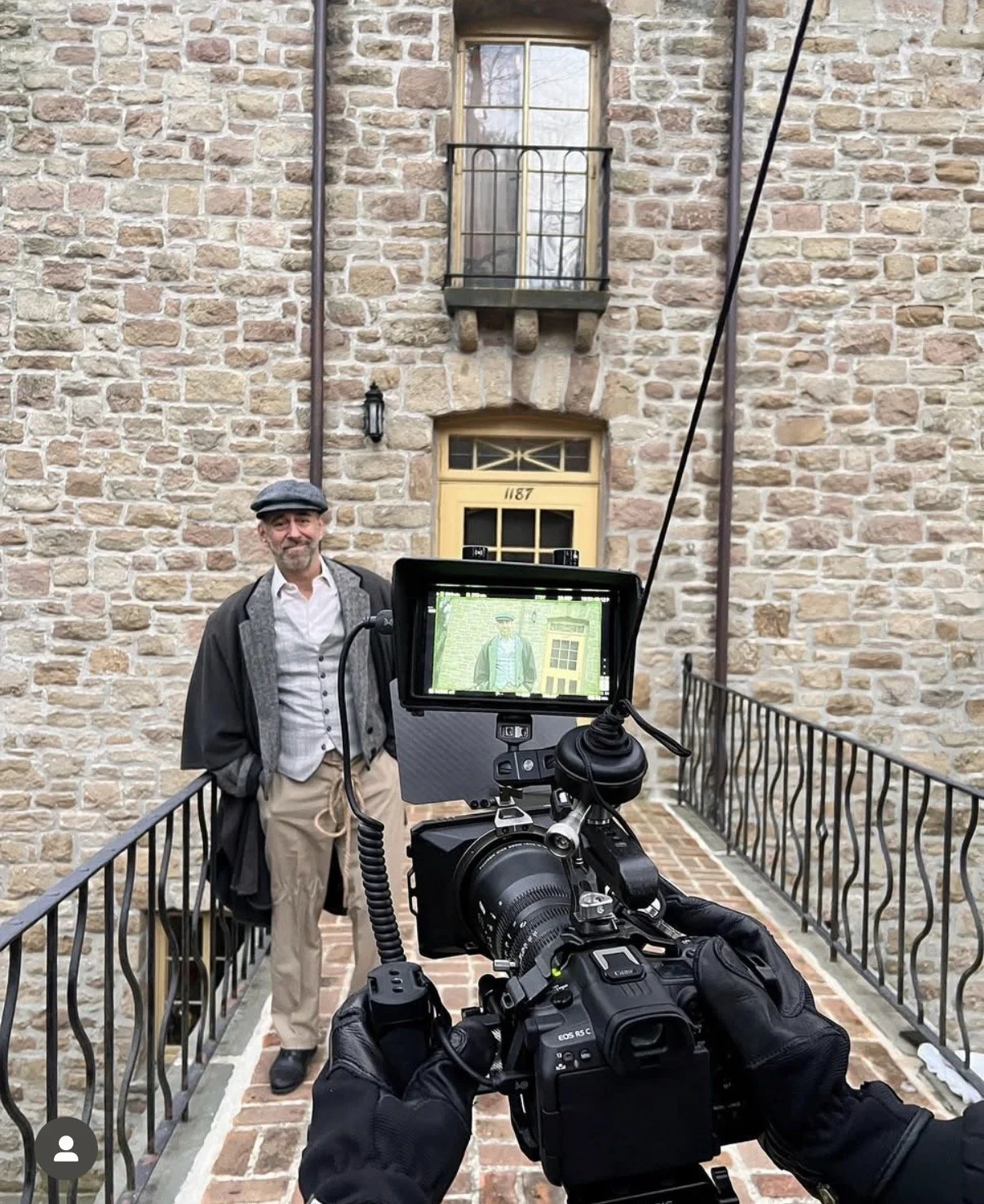A man dressed in period clothing, including a gray cap, a gray vest, and a black coat, stands on a brick walkway in front of a stone building with a yellow door and small balcony. He poses for a photo being taken with a professional camera.