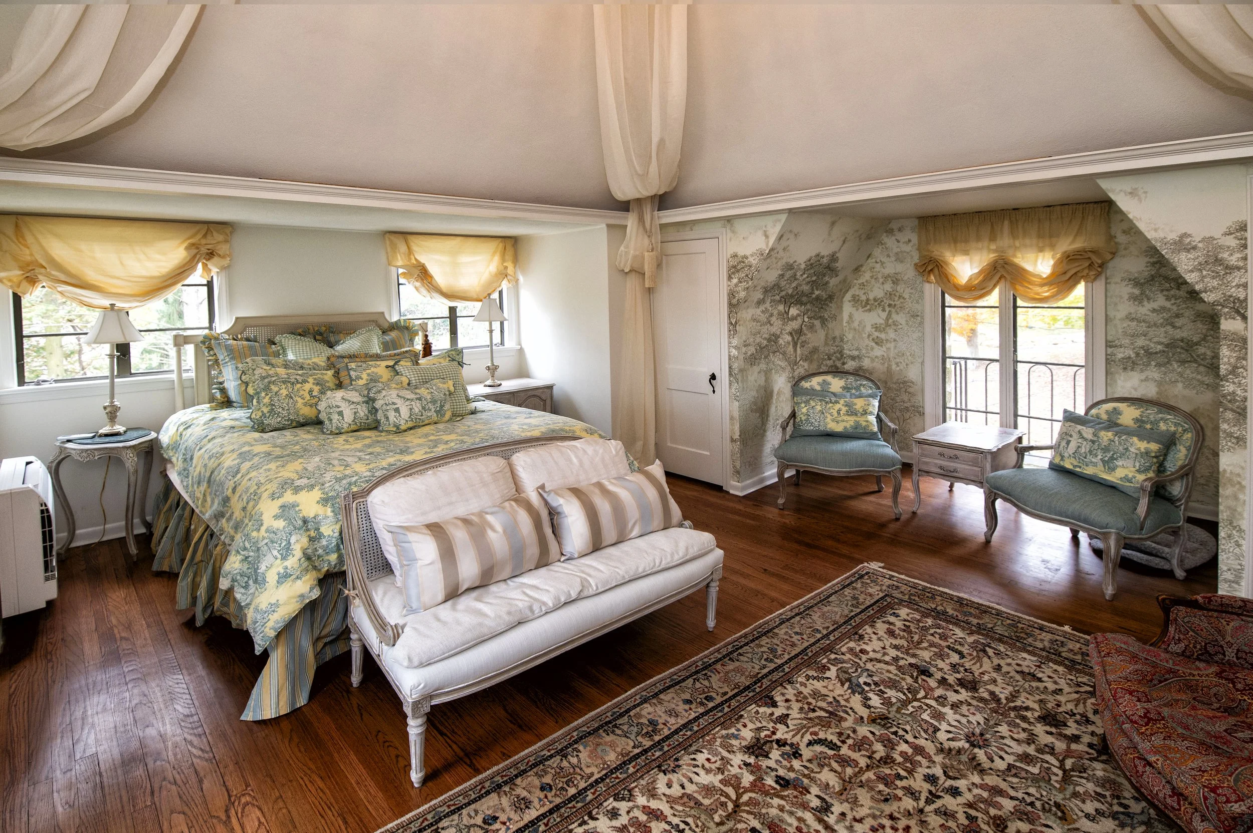 A vintage-style bedroom with a large bed dressed in yellow and blue toile bedding, a white wooden bench at the foot of the bed, two bedside tables with lamps, two windows with yellow curtains, hardwood floors, and a sitting area with two blue tufted armchairs and a small table near a balcony door with a landscape mural wall.