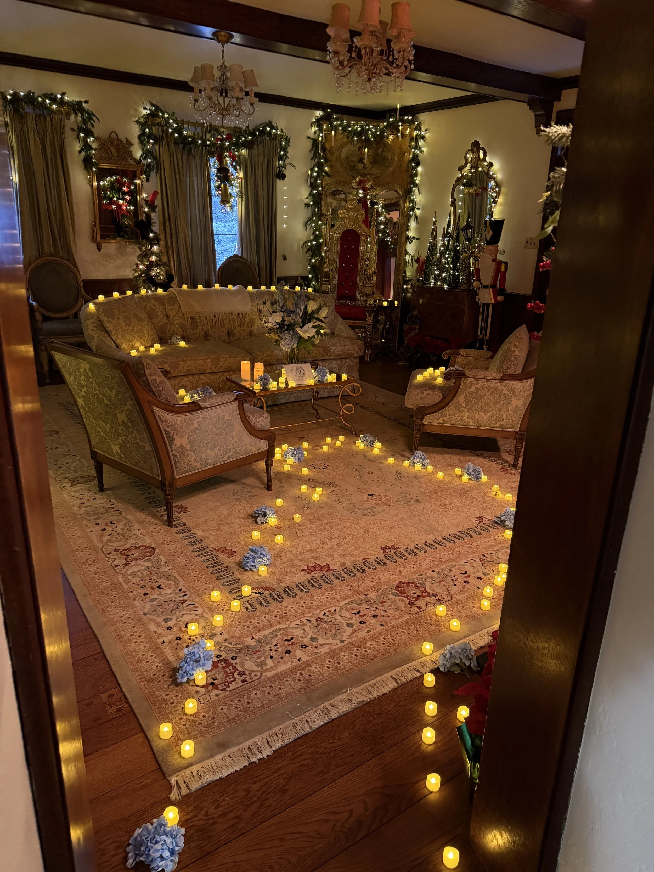 Living room decorated for Christmas with garland and lights, lit candles on furniture and floor, a floral centerpiece table, and an ornate, golden throne-like chair in the corner.