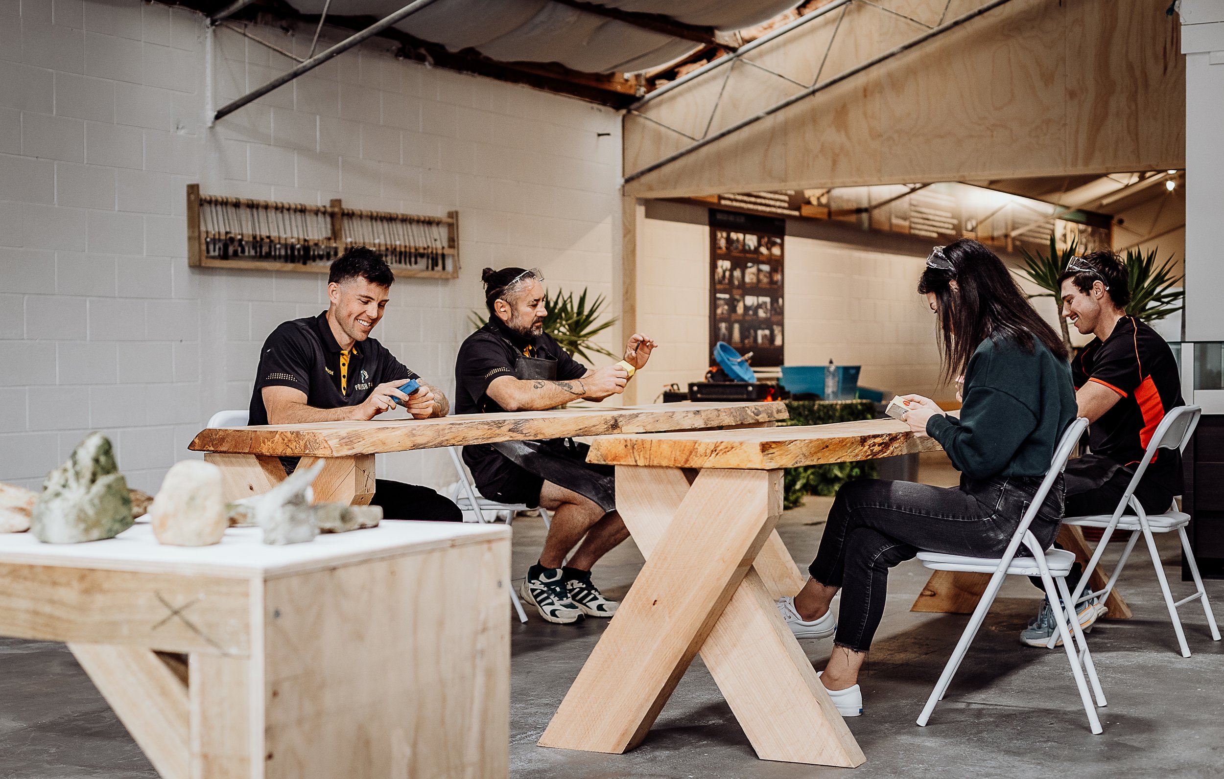 Four people sitting at a wooden table inside a workshop or craft space, smiling and looking at their phones, with rocks on display on a smaller table in the foreground.