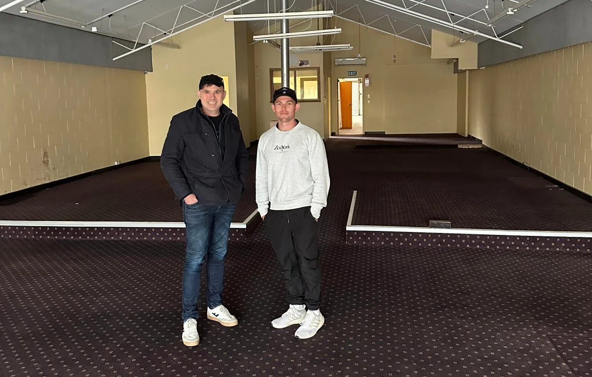 Two men standing inside an empty room with a raised stage and dark patterned carpet, with string lights hanging from the ceiling.