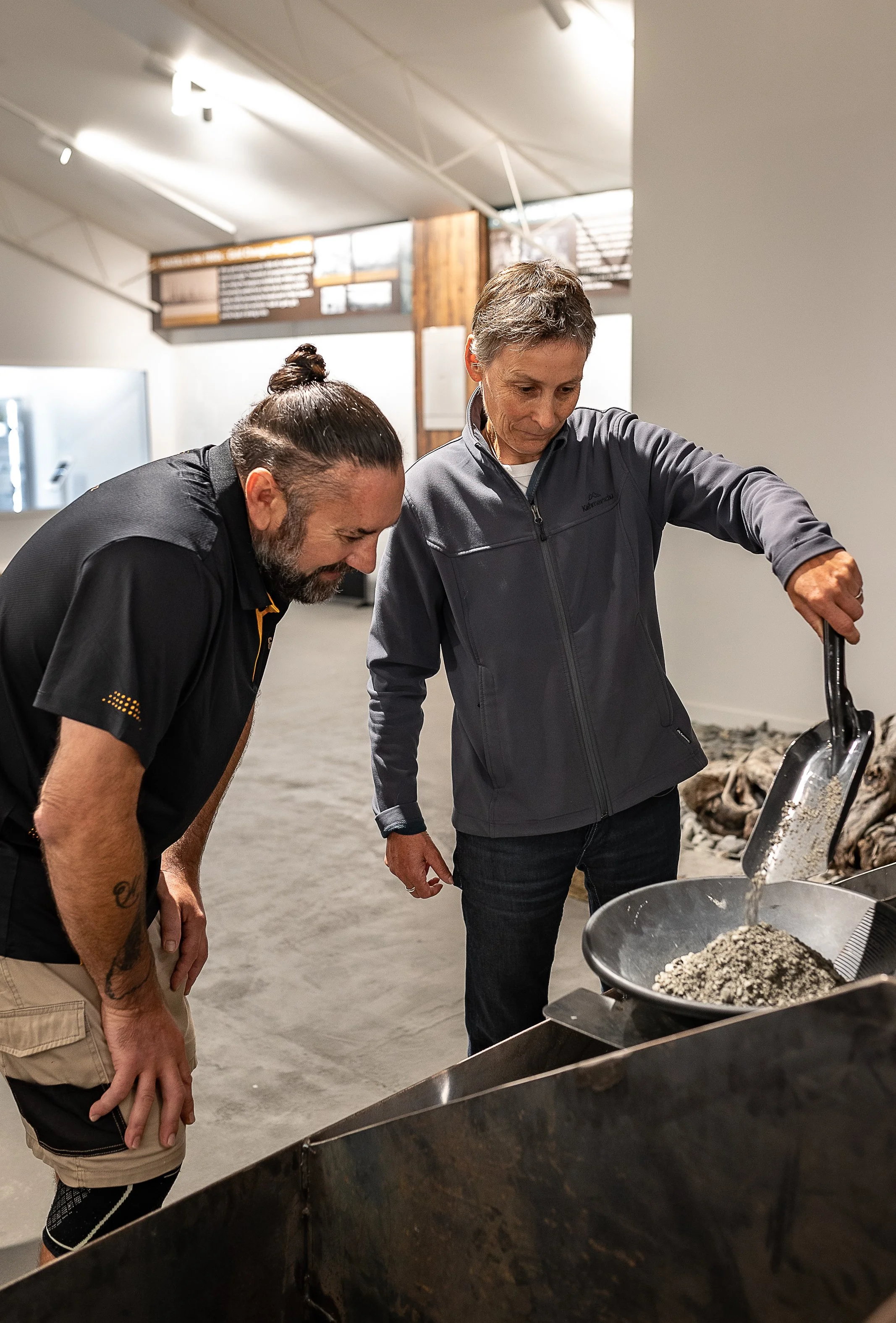 Two men are looking at a large piece of equipment filled with small rocks in an indoor setting with wooden and white walls.