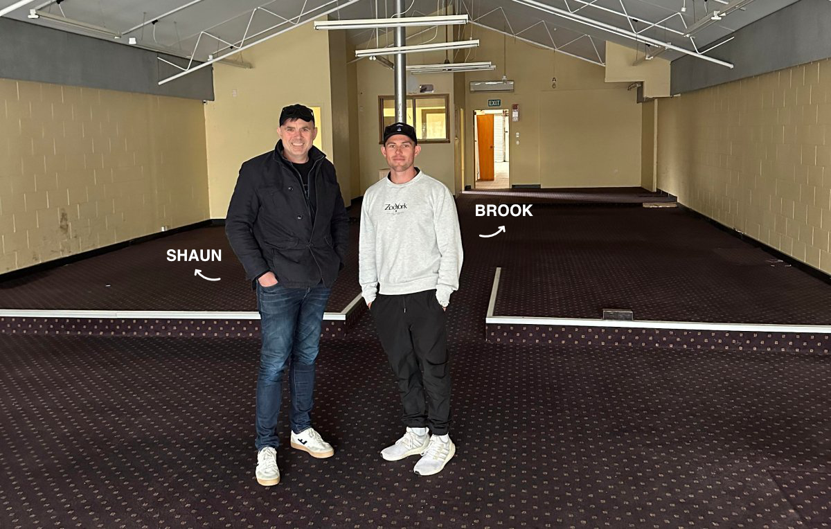 Two men standing in an empty room with a carpeted floor and marked stage area, labeled SHAUN and BROOK, with the room having beige walls and an open door in the background.