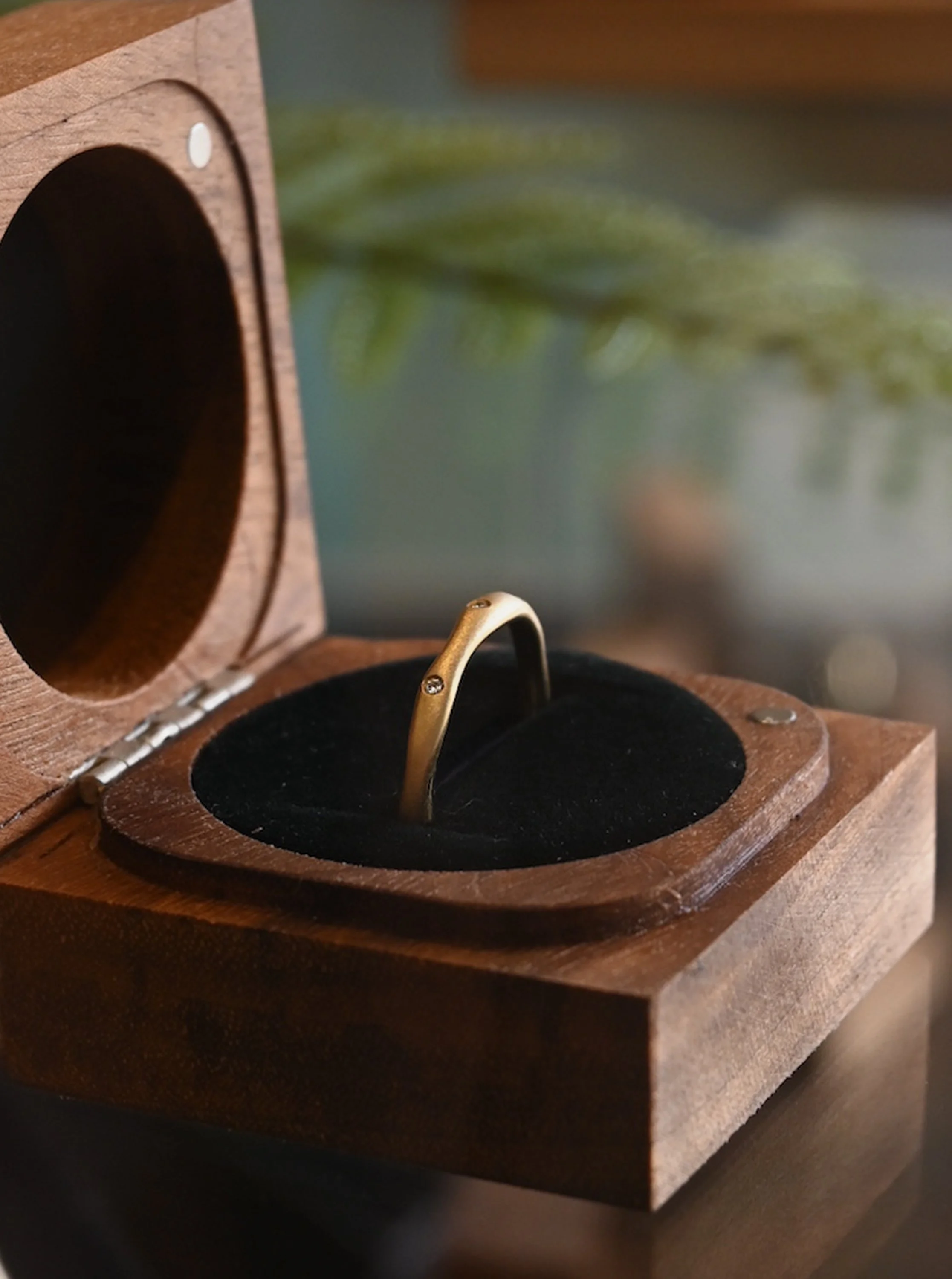 Close-up of a wooden jewelry box with a ring inside. The ring is gold with a small gemstone, placed on a black velvet cushion.