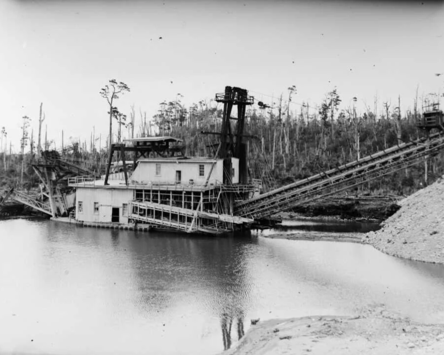 An abandoned gold mining dredge partially submerged in water with trees in the background.