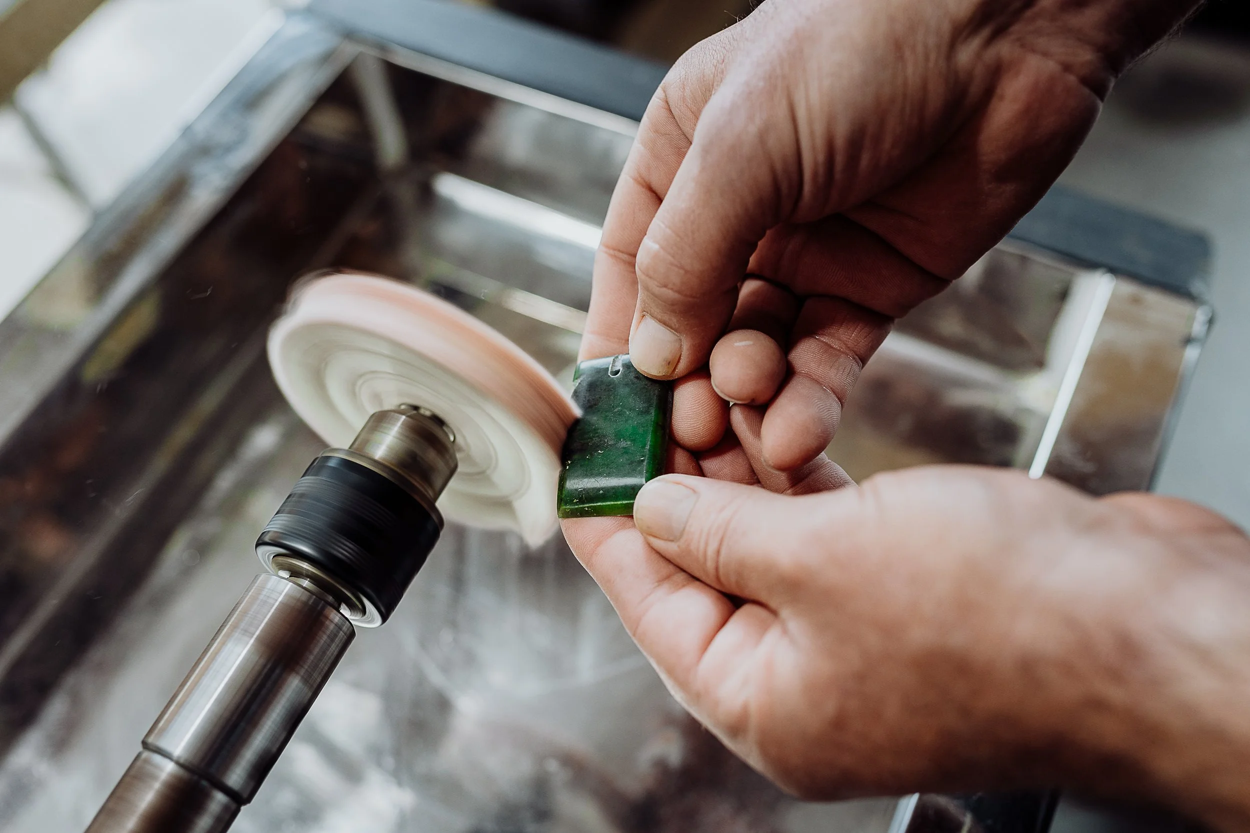 Close-up of a person's hands grinding a piece of green stone on a mechanical polishing wheel.