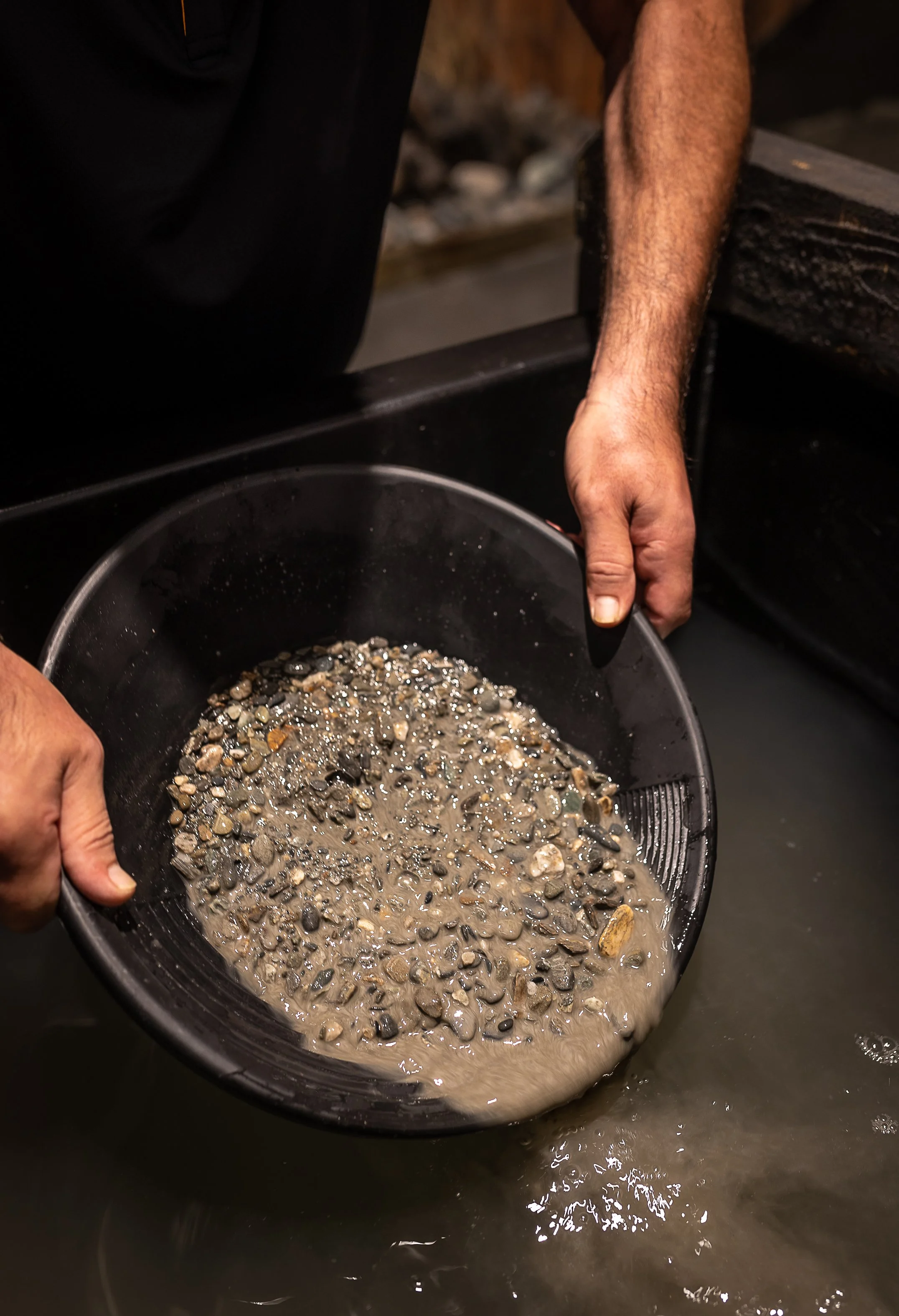 Person holding a black round sieve filled with wet small stones and gravel over a water surface.