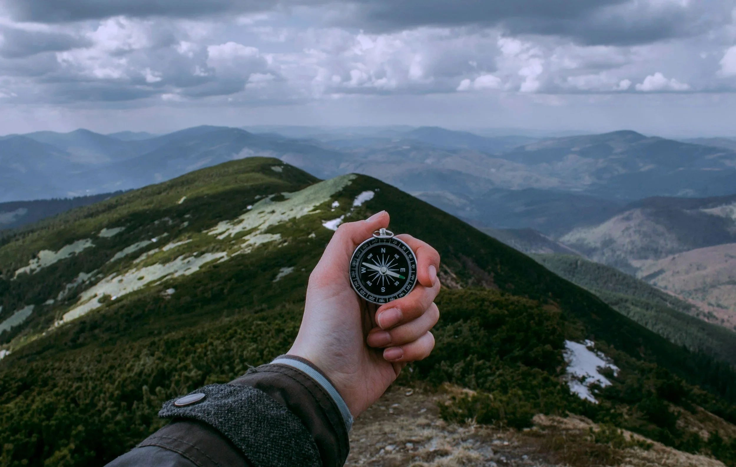 Person holding a compass in front of a mountain range with rolling hills and cloudy sky.