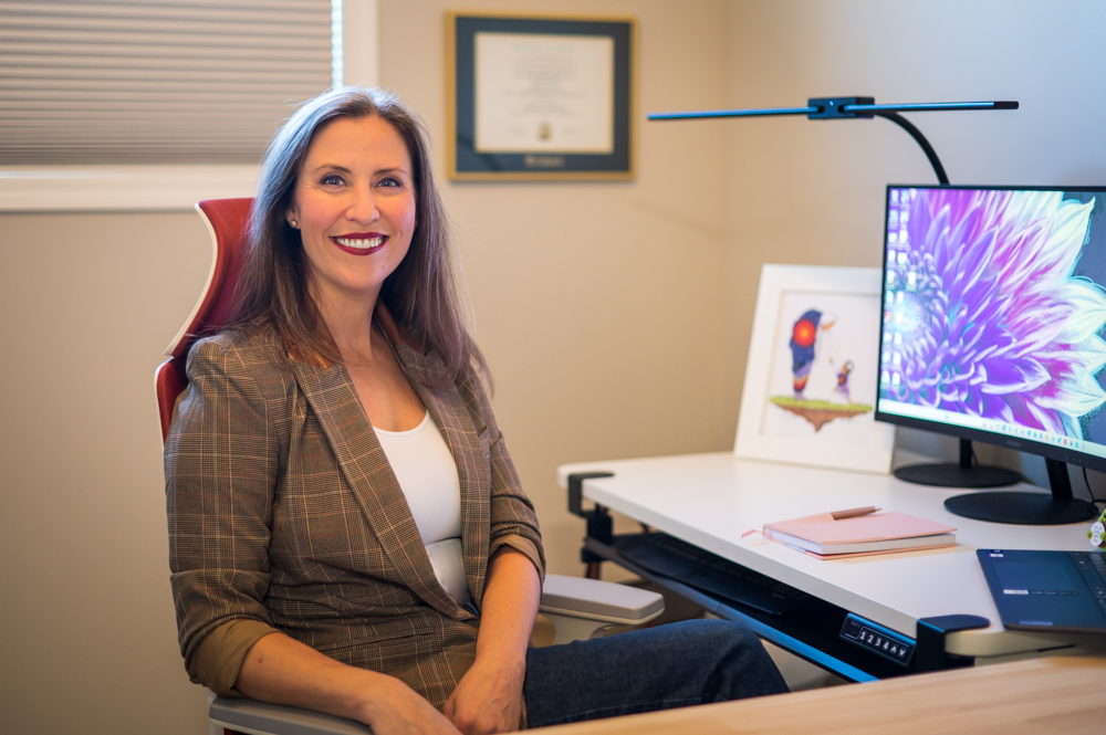 A woman with shoulder-length brown hair and a bright smile sitting in a home office. She is wearing a plaid blazer over a white top. Behind her are a framed certificate, a window with blinds, and a desk with a computer monitor, a pink notebook, a drawing frame with artwork, and other office supplies.