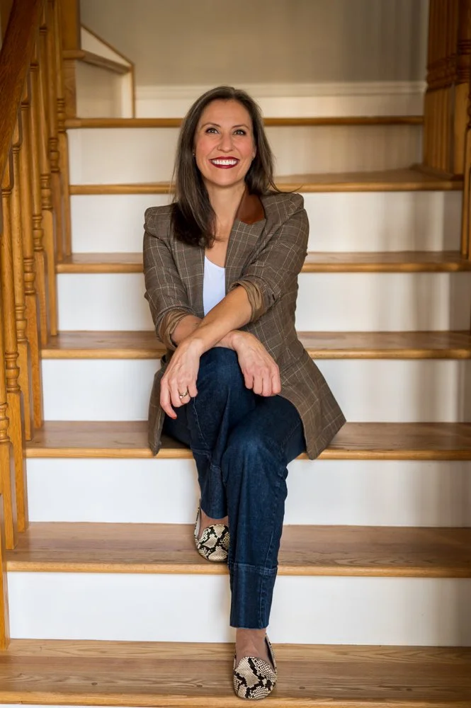 A woman with long dark hair and red lipstick sitting on wooden stairs, smiling, wearing a brown plaid blazer, white top, blue jeans, and snakeskin print shoes.