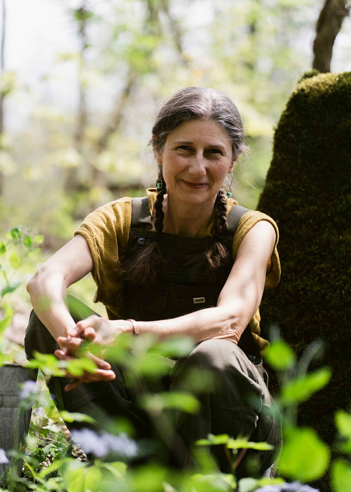 An older woman with braided hair, wearing a yellow top and black overalls, sitting outdoors in a forested area, smiling at the camera.