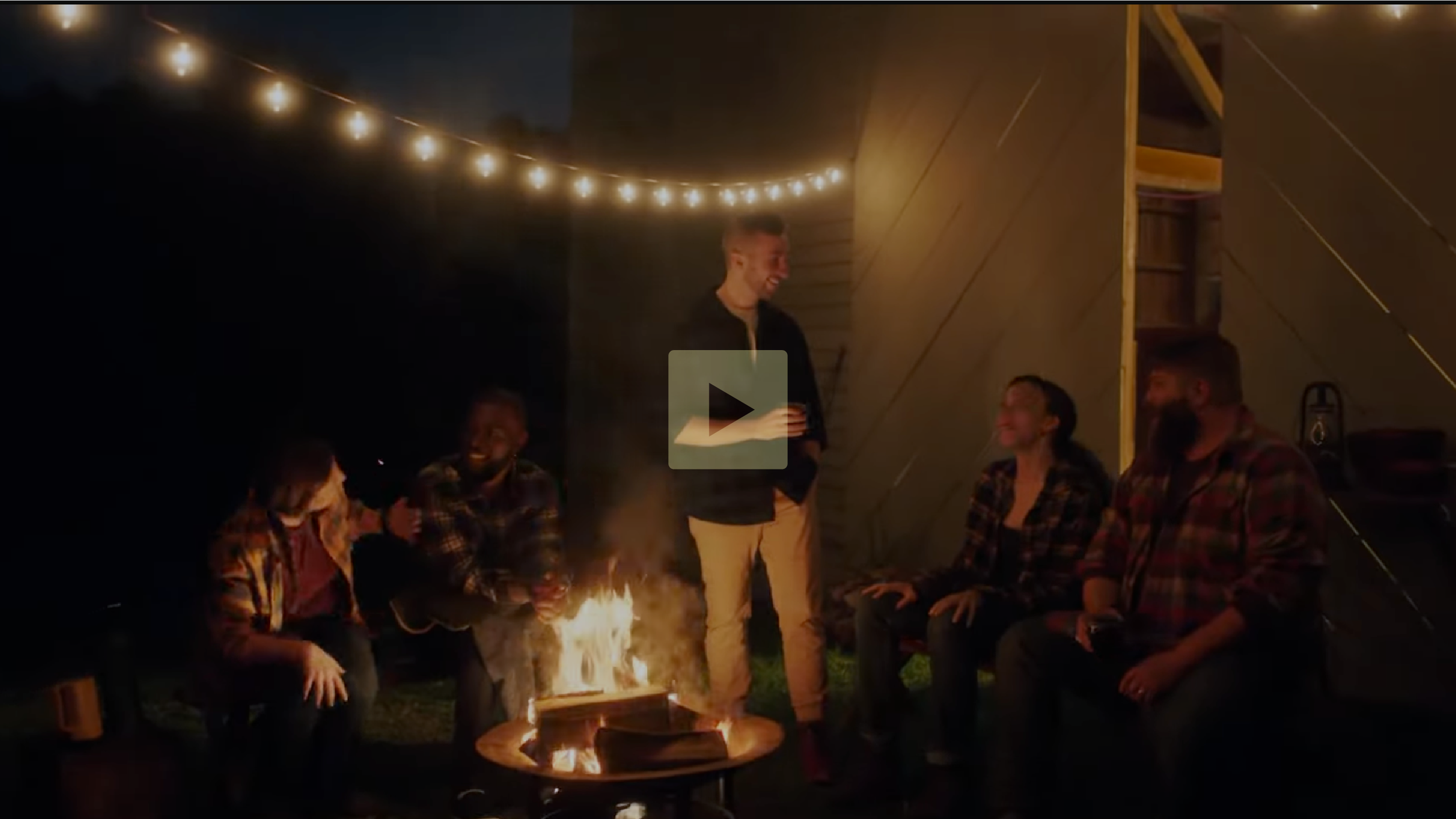 Group of five friends sitting and standing around a fire pit outdoors at night, illuminated by string lights overhead, smiling and chatting.