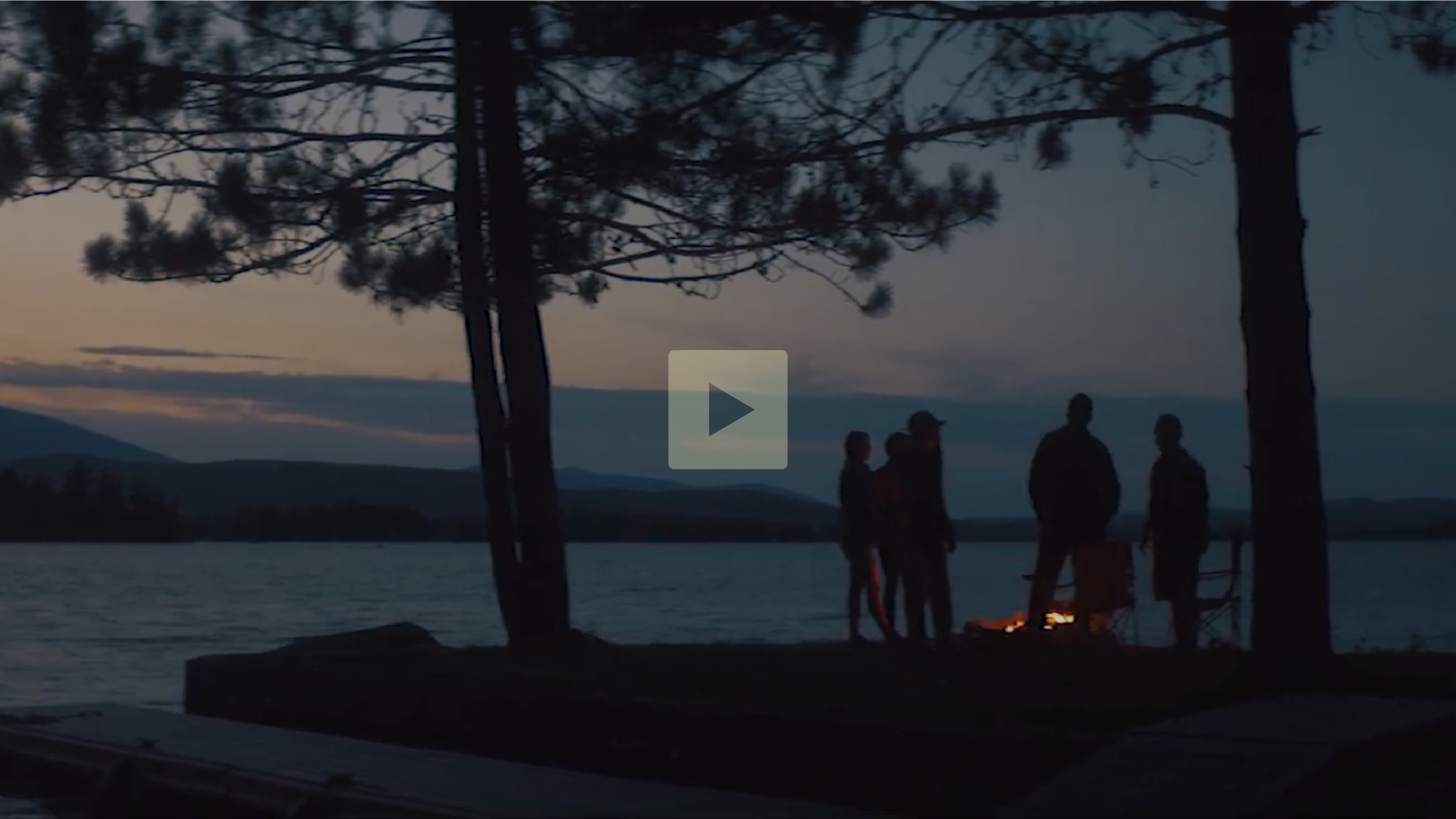A group of five people standing around a campfire by a lake at dusk, with trees and mountains in the background.