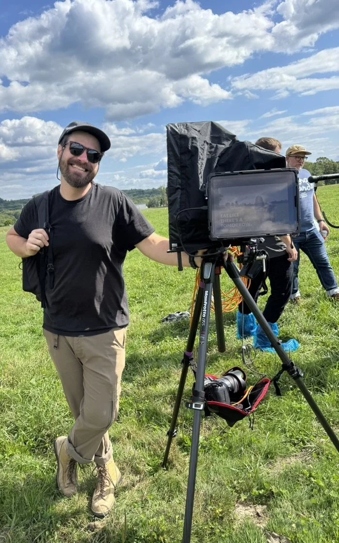 A smiling man with sunglasses, a black T-shirt, beige pants, and hiking boots standing outdoors next to a large camera on a tripod. He holds a strap over his shoulder. In the background, there are two other people, a man and a woman, walking across a