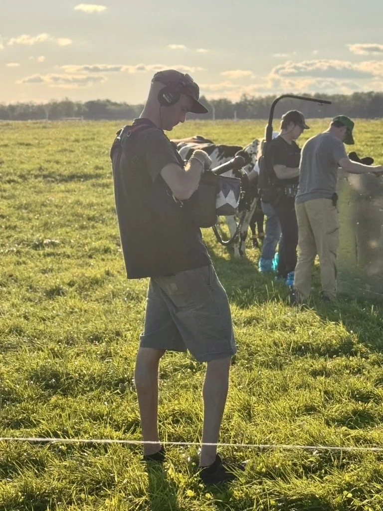 Young man standing on grass at outdoor event, using a camera or equipment, with others in line in the background, under a partly cloudy sky during late afternoon or evening.