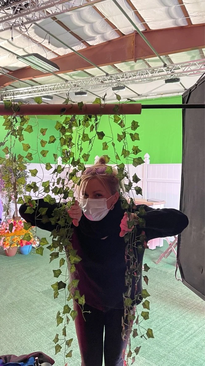 A woman in a face mask and black clothing hiding behind hanging leaves and vines, with a green screen and studio equipment visible in the background.