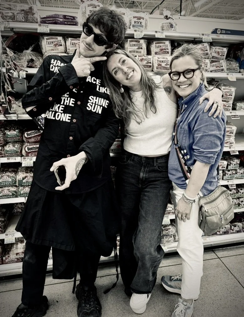 Three friends standing together inside a grocery store aisle, posing happily for a photo. The background shows shelves stocked with various bags of snacks and food items.