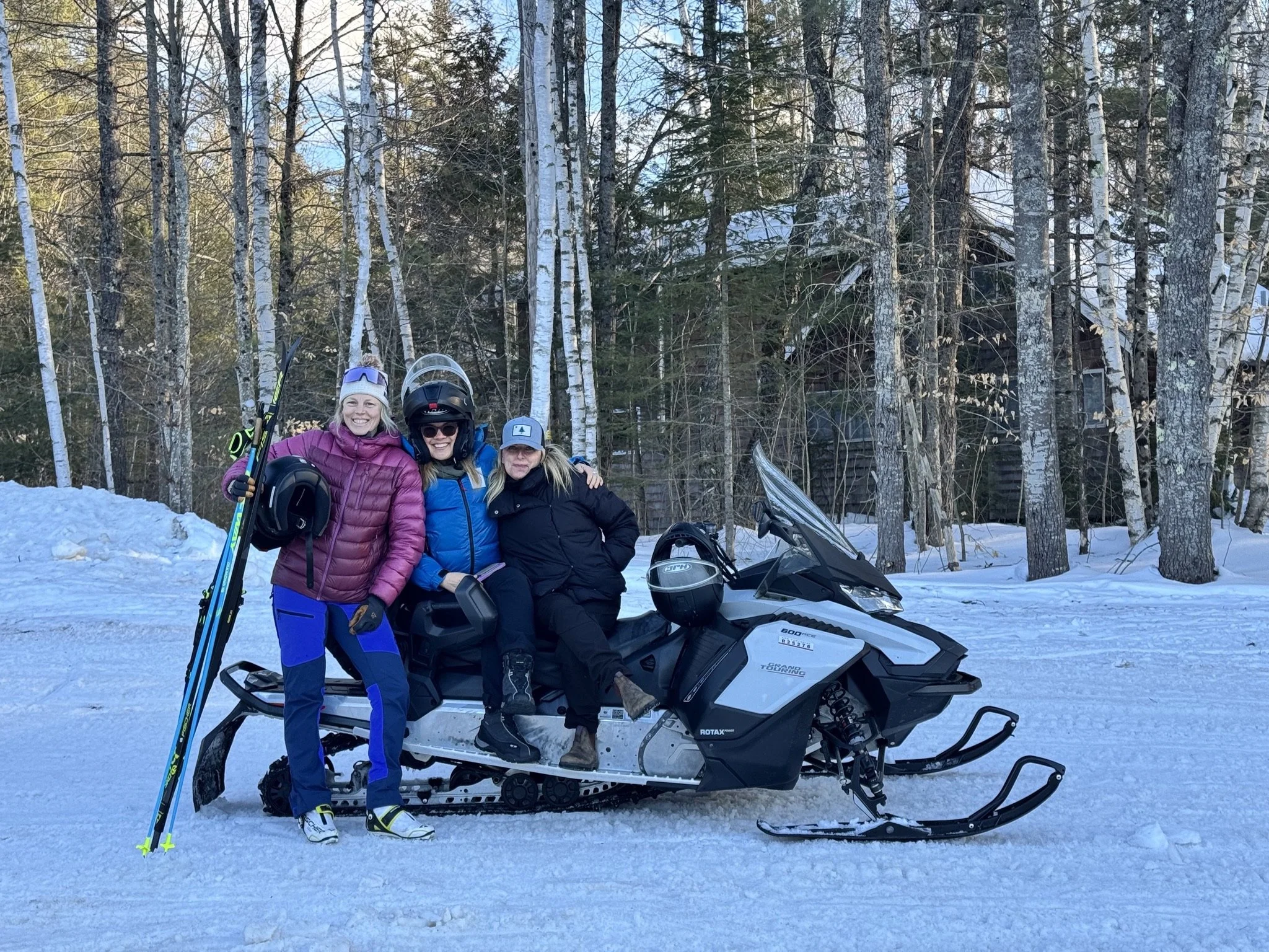 Three women in winter gear with ski poles and a helmet, sitting on a snowmobile in a snowy forest with bare trees and a building in the background.