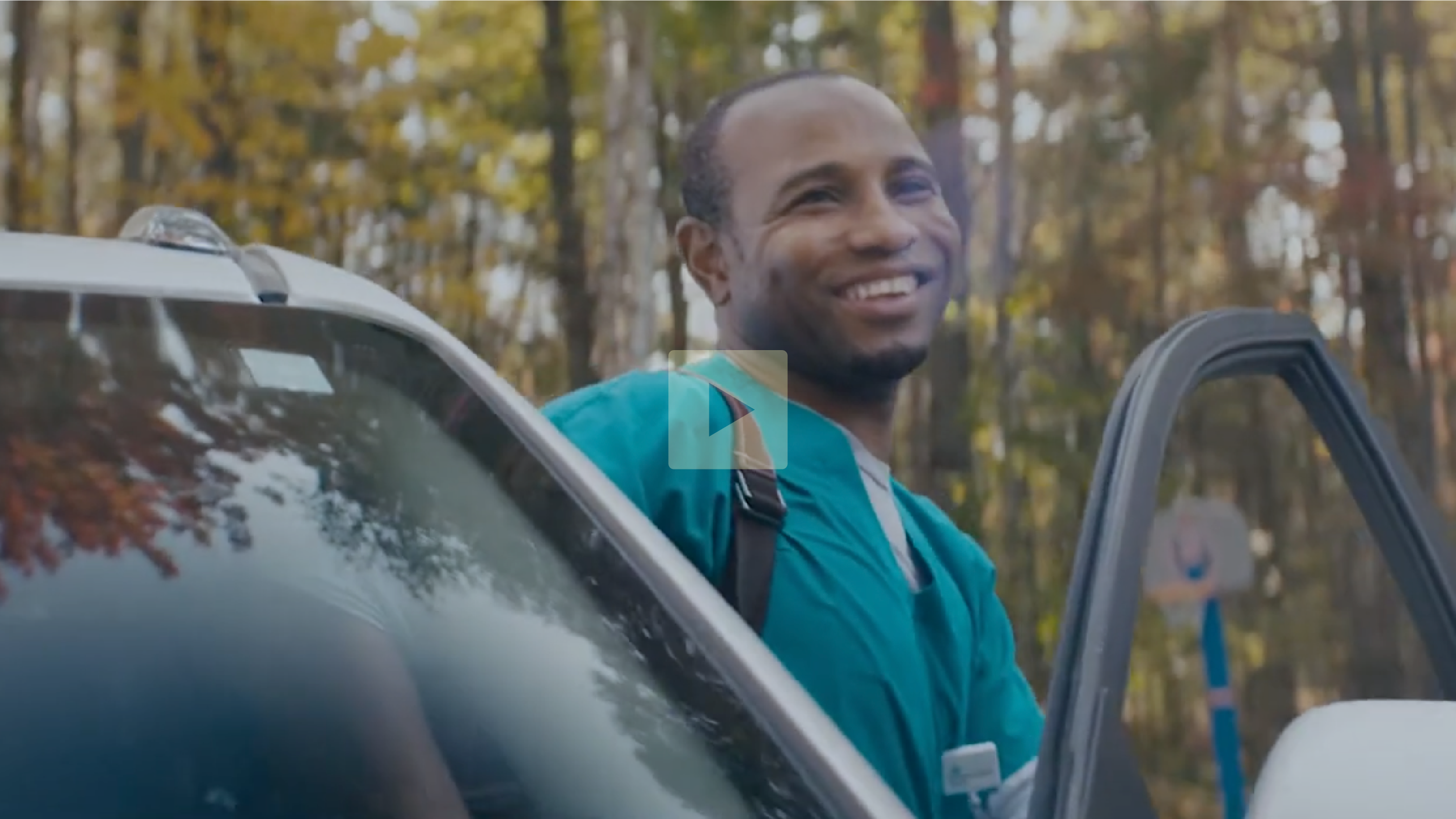 A man smiling while standing next to a white car with an open door in a wooded area with trees showing autumn foliage.