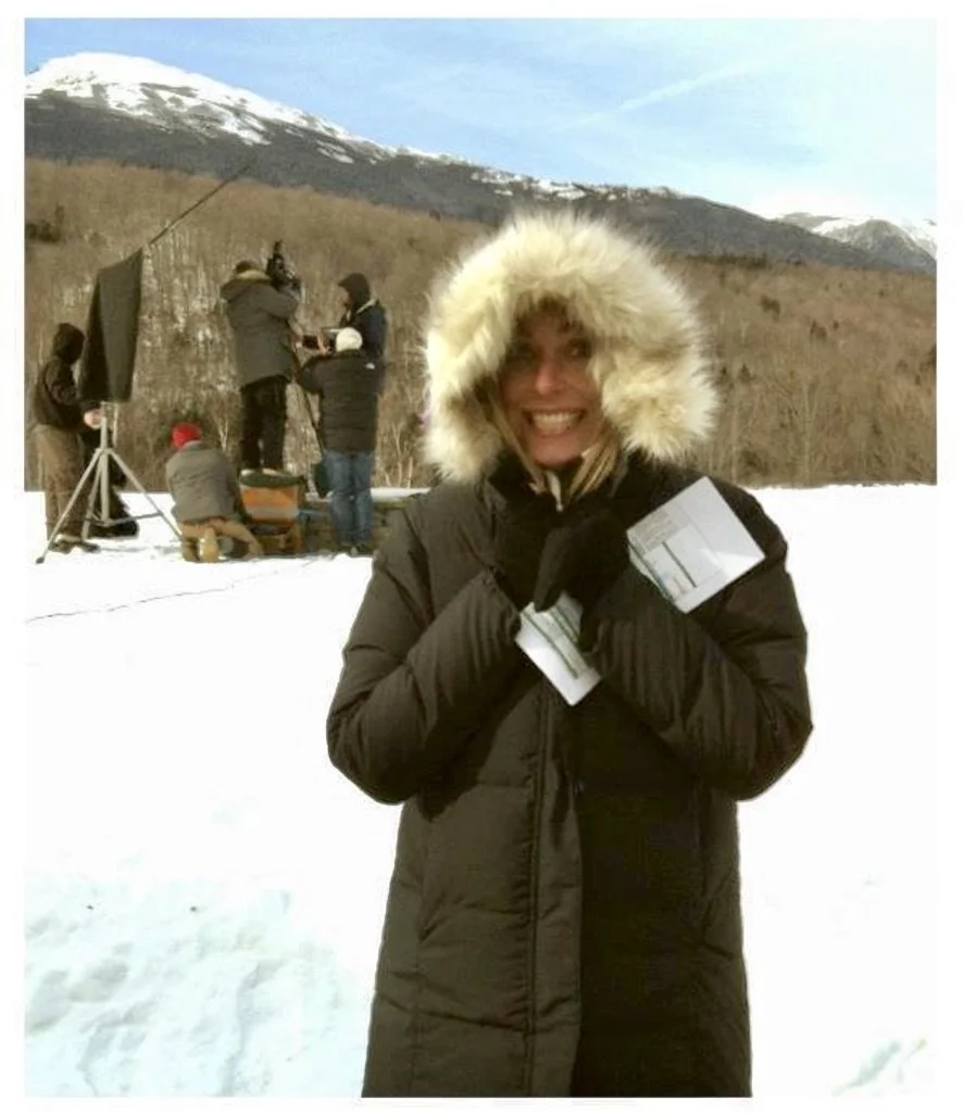 A woman wearing a black winter coat with a fur-lined hood, smiling and holding papers, standing in a snowy landscape during a film or photo shoot with mountains in the background.